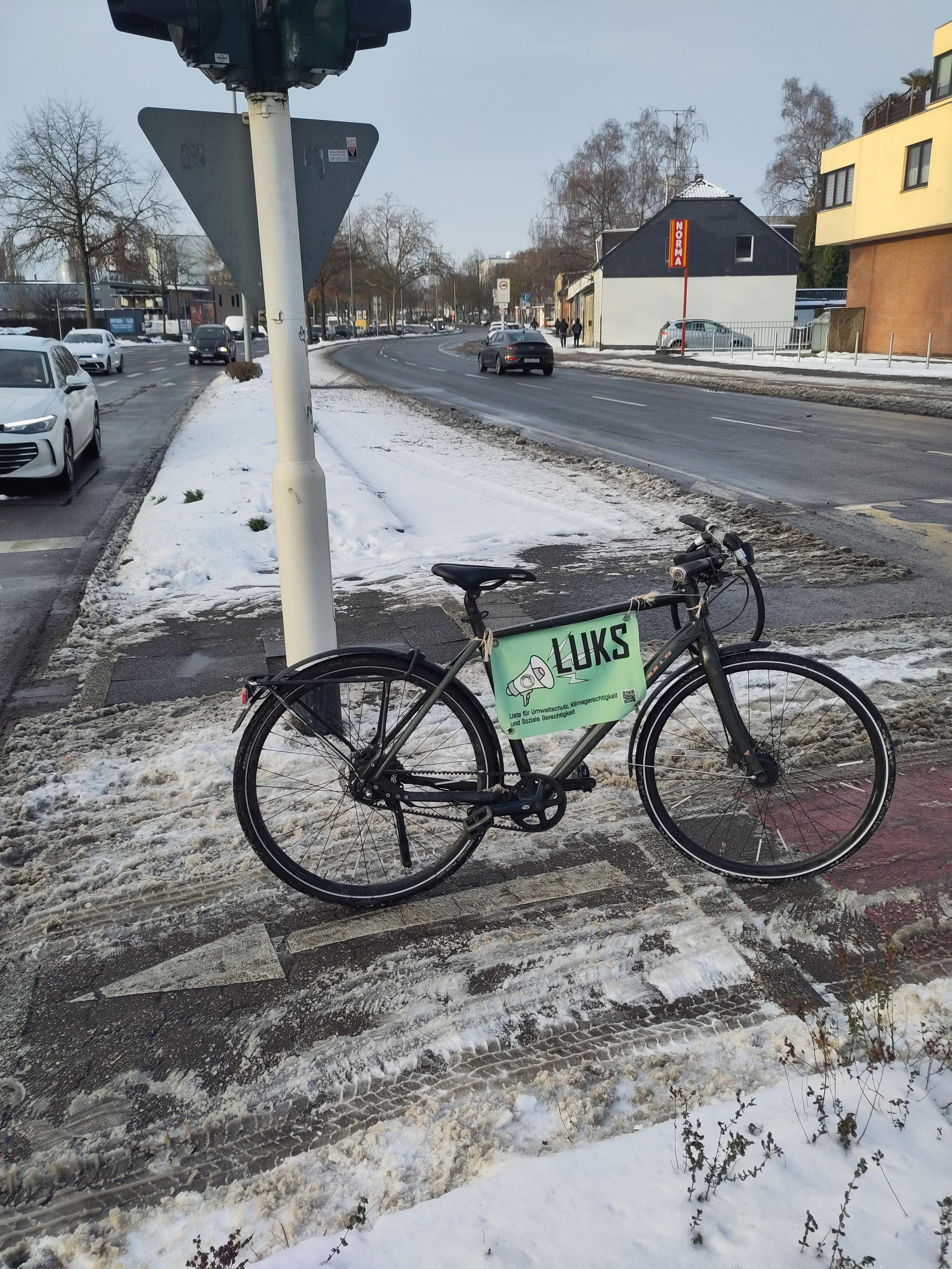 Man sieht ein Fahrrad, das auf einer (z.B. für Lastenräder, Fahrrad mit Kinderanhänger ) viel zu schmalen Verkehrsinsel steht. Einseitig neben der Verkehrsinsel zeigt weißer, unbefahrener Schnee, dass die Autos diesen Platz nicht einmal benötigen und nach dorthin die Verkehrsinsel einfach erweitert werden könnte.