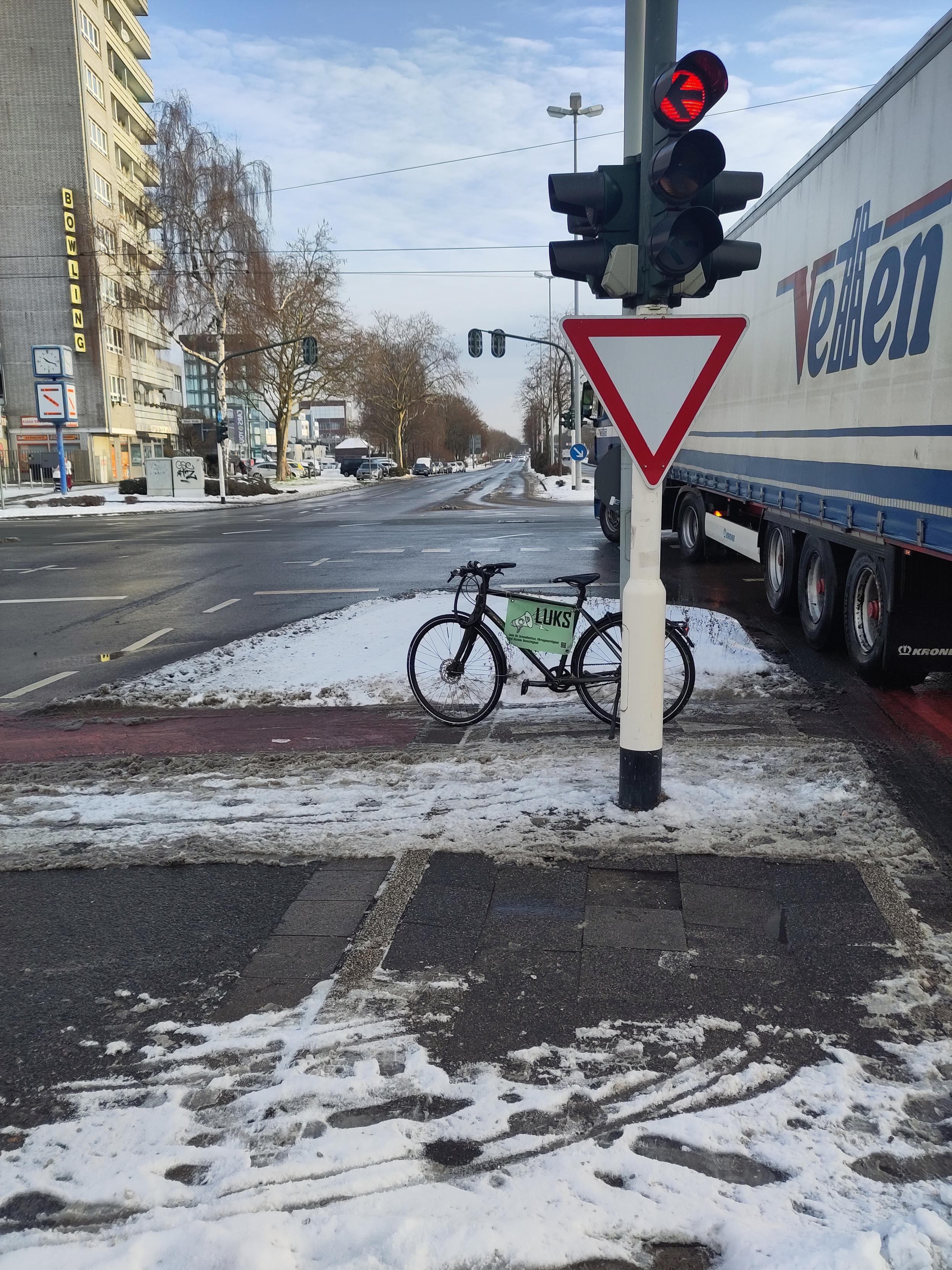 Man sieht ein Fahrrad, das auf einer (z.B. für Lastenräder, Fahrrad mit Kinderanhänger ) viel zu schmalen Verkehrsinsel steht. Einseitig neben der Verkehrsinsel zeigt weißer, unbefahrener Schnee, dass die Autos diesen Platz nicht einmal benötigen und nach dorthin die Verkehrsinsel einfach erweitert werden könnte.