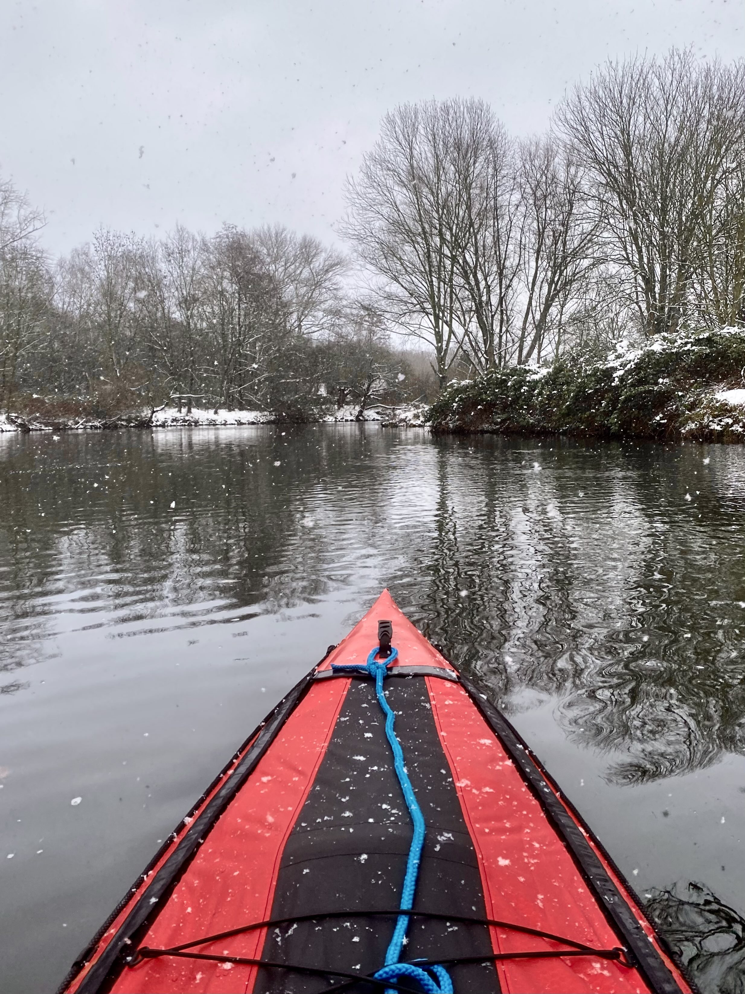 Ein ruhiger Fluss aus der Perspektive des Kayakfahrers. Der Bug des schwarz-roten Schlauchkajaks ragt vom unteren Rand bis in die Bildmitte hinein. Es schneit leicht, die Ufer sind mit Schnee bedeckt. Die blätterlosen Bäume spiegeln sich im Wasser, der Himmel ist bedeckt und eintönig grau.
