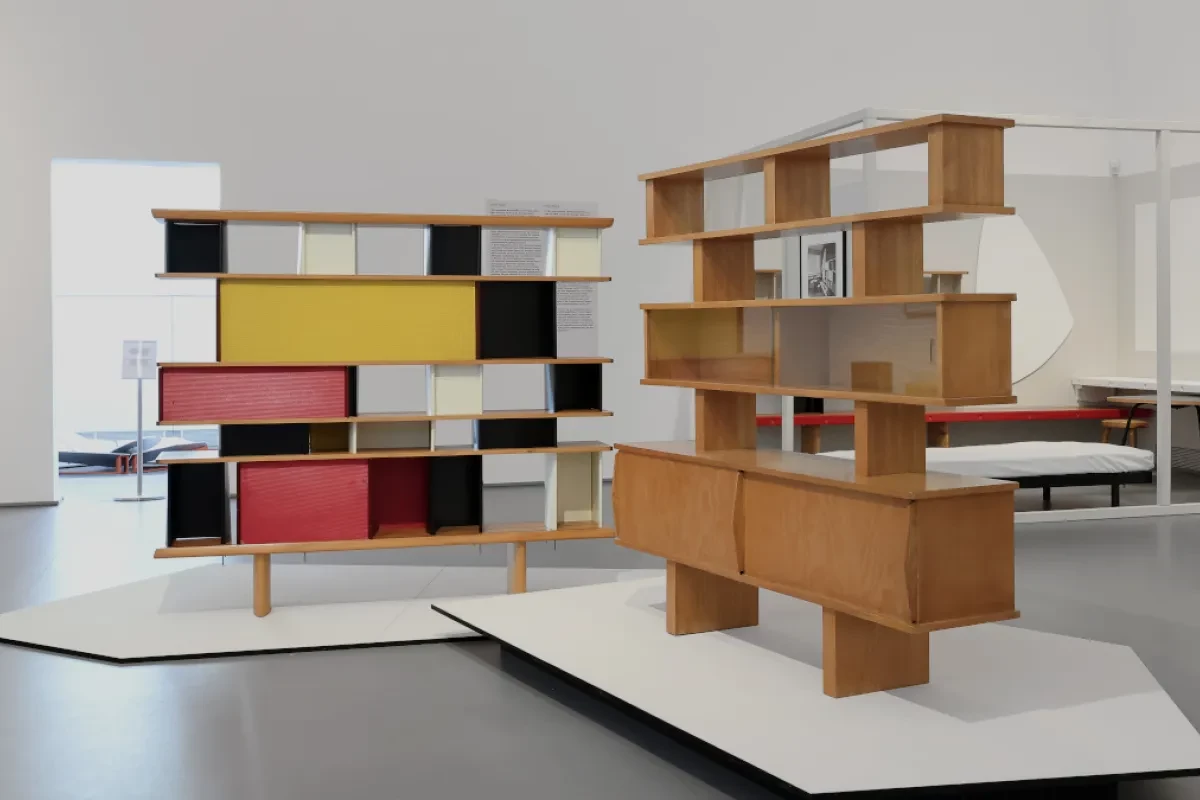 Zwei modernistische Regalsysteme als Ausstellungsstücke in einem hellen Museumsraum: links ein farbiges Regal mit gelben, roten, weißen und schwarzen Flächen, rechts ein offenes Regal aus hellem Holz mit integrierten Schrankelementen. Beide Möbel stehen auf weißen Podesten, im Hintergrund sind weitere Möbel und Ausstellungsbereiche zu sehen.