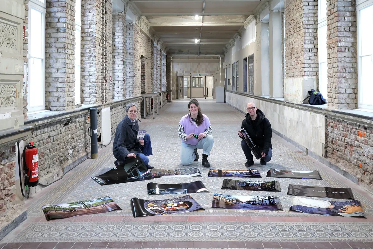 Drei Personen knien in einem langen Flur eines offenbar renovierten Gebäudes mit freigelegten Backsteinpfeilern, großen Fenstern und gemustertem Fliesenboden. Vor ihnen liegen mehrere großformatige Fotodrucke und Plakate ausgebreitet; eine Person hält Prospekte, eine andere einen gerollten Druck. Links ist ein Feuerlöscher an der Wand montiert, im Hintergrund führt der Flur zu einer Tür unter Deckenlampen.