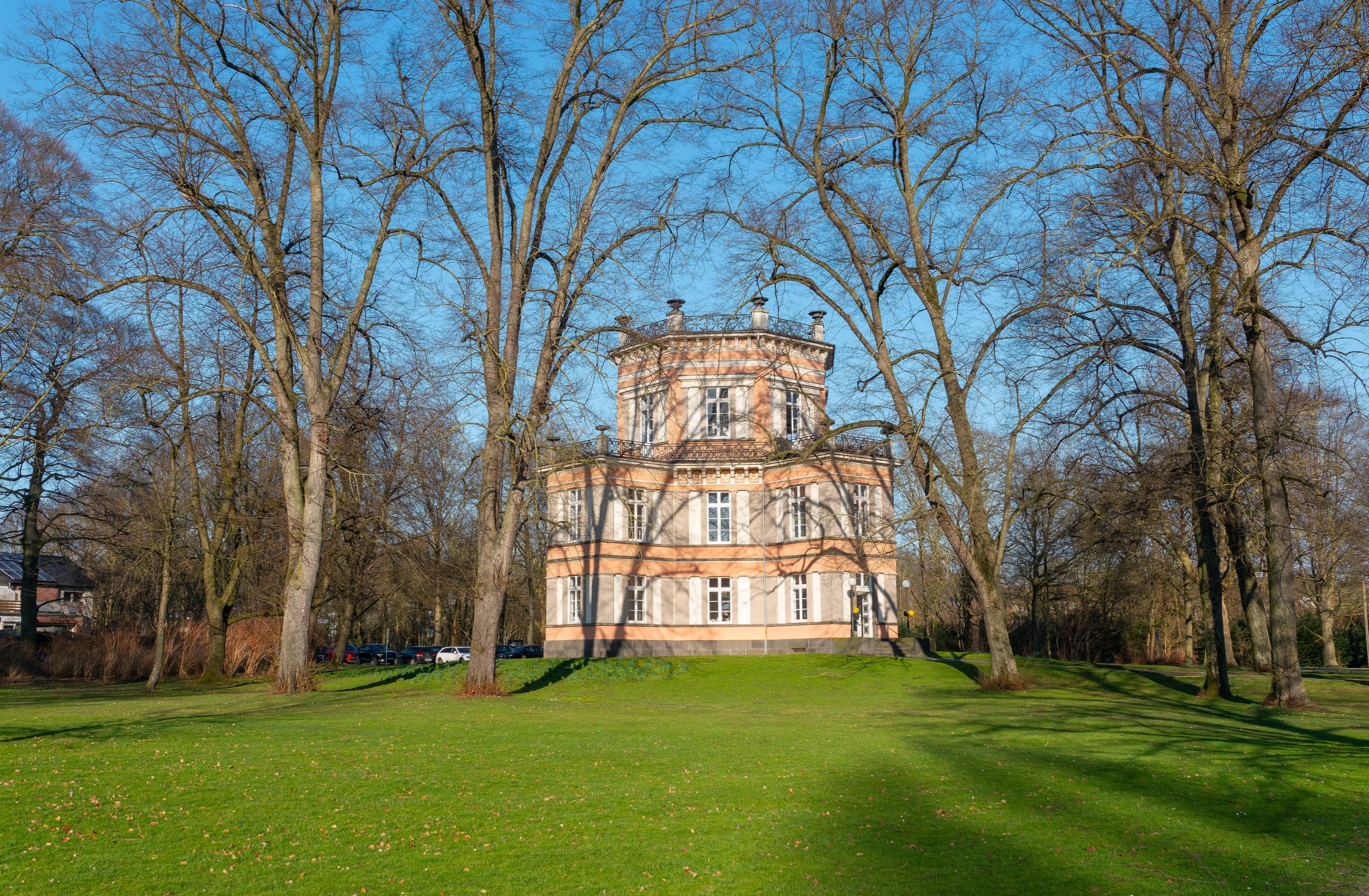 Historische, hellrosa-weiße Villa in einem weitläufigen Park, mittig auf einer grünen Wiese gelegen. Das mehrgeschossige Gebäude mit achteckigem Obergeschoss ist von hohen, noch unbelaubten Bäumen umgeben. Über der Szene spannt sich ein klarer, leuchtend blauer Himmel, während lange Schatten auf das Gras fallen.
