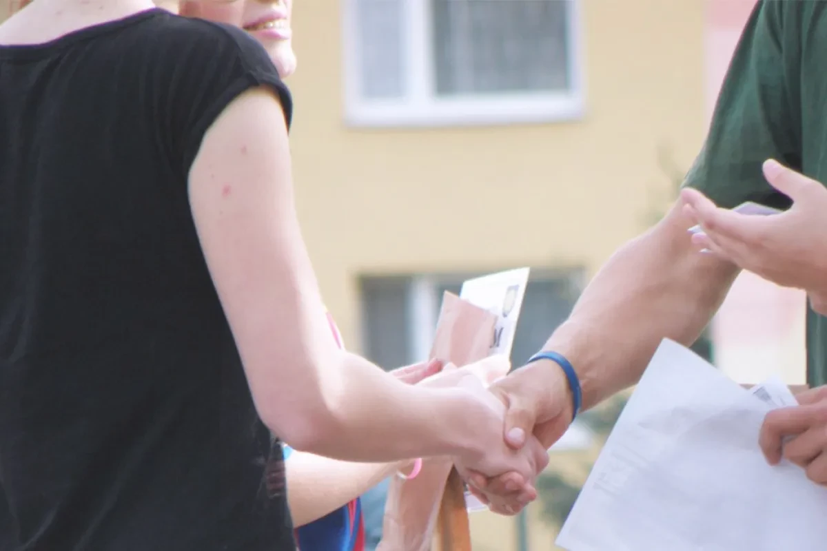 Nahaufnahme von zwei Menschen, die sich im Freien die Hände schütteln. Eine Person hält dabei Papiere in der Hand, im Hintergrund ist unscharf die Fassade eines Gebäudes zu sehen.