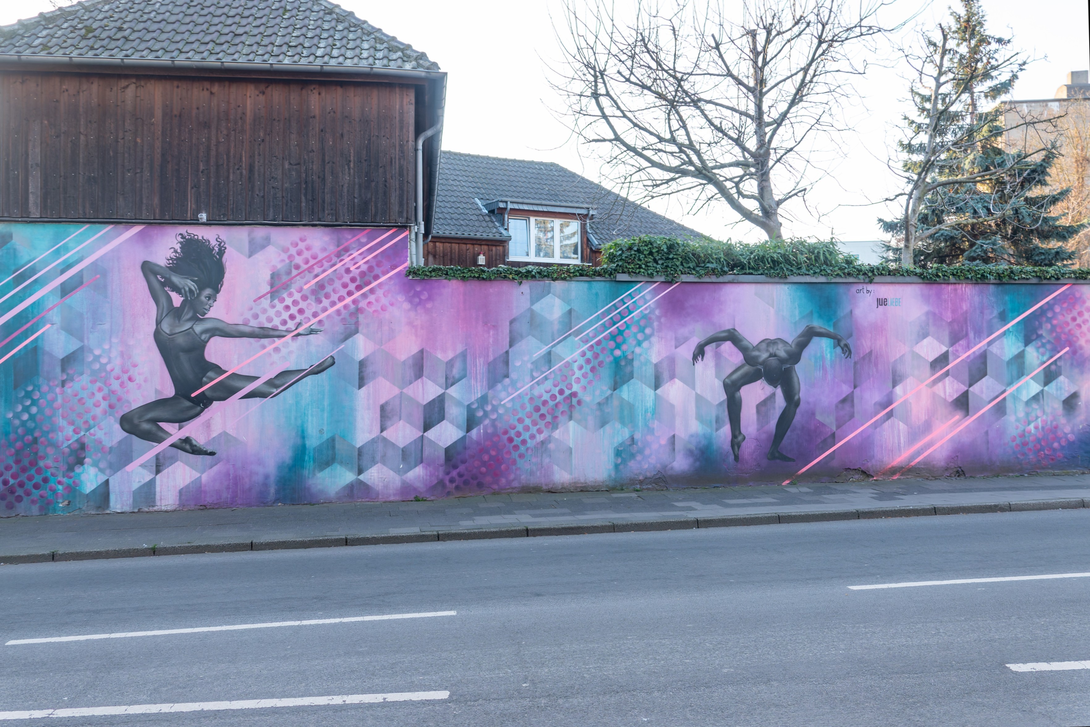 Großflächiges Streetart-Mural an einer langen Mauer entlang einer Straße. Das Bild ist in Violett-, Rosa- und Türkistönen gehalten und zeigt zwei schwarzgraue, tänzerisch wirkende menschliche Figuren vor einem geometrischen Hintergrund aus Hexagonen, Punkten und schrägen Neonlinien. Links springt eine Figur dynamisch mit ausgestrecktem Arm, rechts ist eine zweite Figur in einer gebückten, fast akrobatischen Pose zu sehen. Hinter der bemalten Mauer stehen ein dunkles Holzhaus, kahle Bäume und eine Tanne. Im Vordergrund verläuft eine breite, leere Fahrbahn.