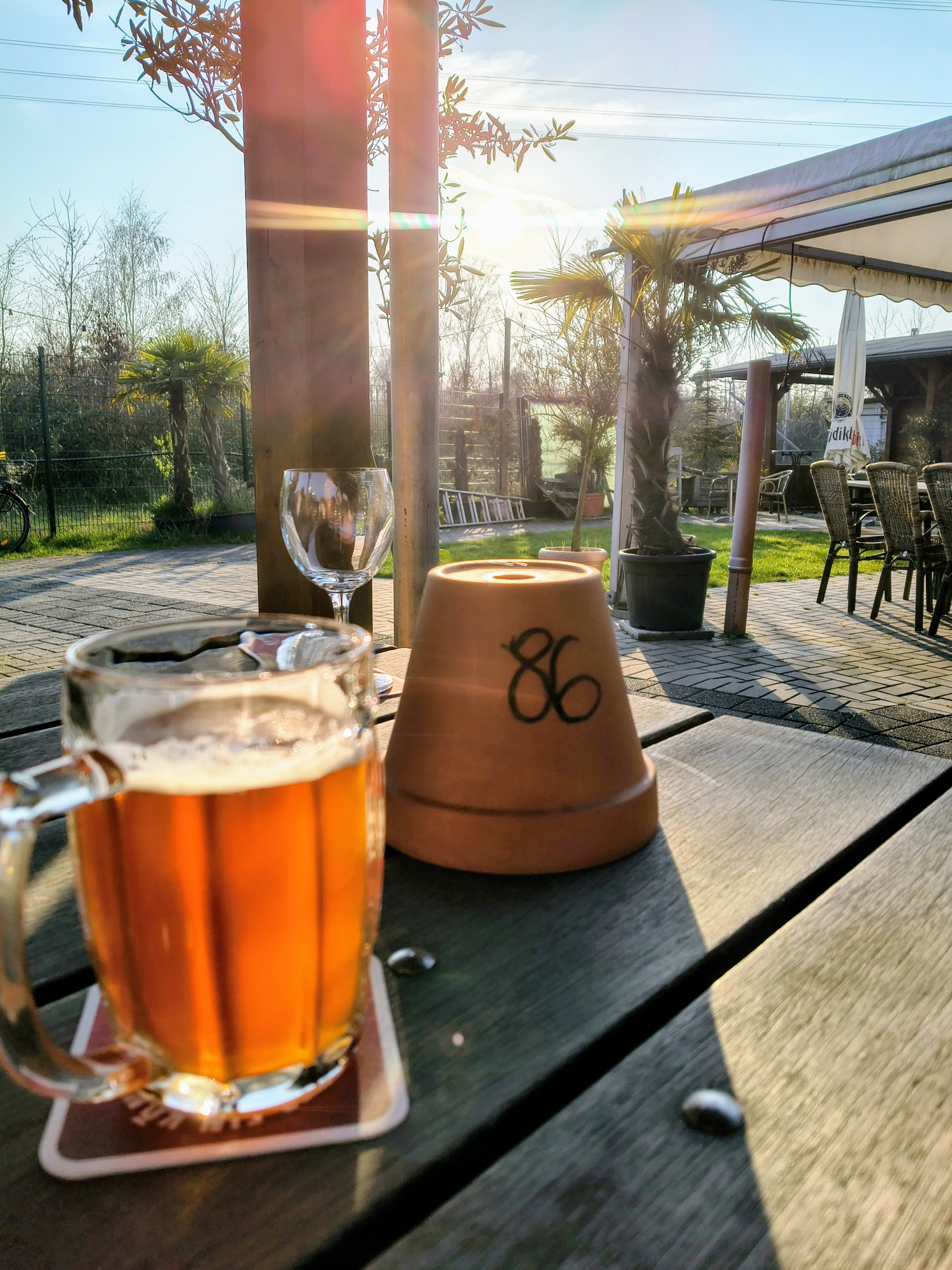 Eine herbstliche Außenaufnahme in einer Gastronomie bei tiefstehender Abendsonne. Im Vordergrund steht auf einem dunklen Holztisch ein gefüllter Glasbierkrug mit bernsteinfarbenem Bier und weißer Schaumkrone auf einem Untersetzer. Daneben steht ein umgedrehter Terrakotta-Blumentopf, auf dem die Zahl „86“ mit schwarzem Marker steht. Im Hintergrund ist ein leeres Weinglas zu sehen.
​Die Szenerie wird von links durch starkes Gegenlicht beleuchtet, das rötliche Lichtreflexe (Lens Flares) erzeugt. Der Außenbereich ist mit hellgrauem Kopfsteinpflaster ausgelegt, auf dem lange Schatten fallen. Im Mittelgrund stehen dunkle Korbstühle unter einer weißen Markise und eine kleine Palme im Topf. Der Hintergrund zeigt einen Garten mit Rasen, einem Maschendrahtzaun und entlaubten Bäumen unter einem hellen, klaren Himmel mit fernen Stromleitungen.