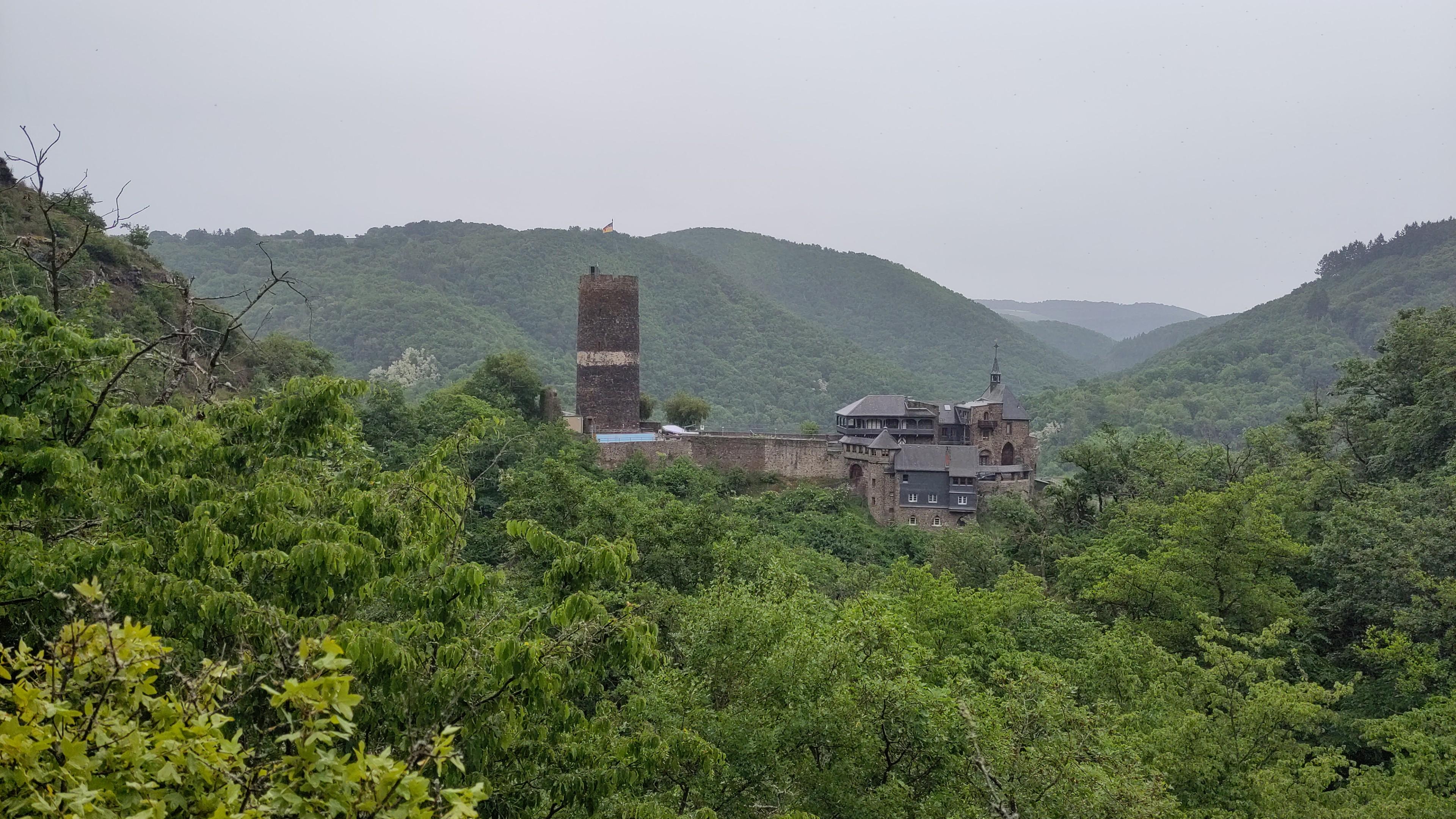 Zwischen bewaldeten Berghängen liegt eine braun-graue Burg. Rechts ein Gebäudekomplex mit grauen Schieferdächern und einem Kapelltürmchen, links ein hoher Bergfried mit einem hellen umlaufenden Putzstreifen auf halber Höhe