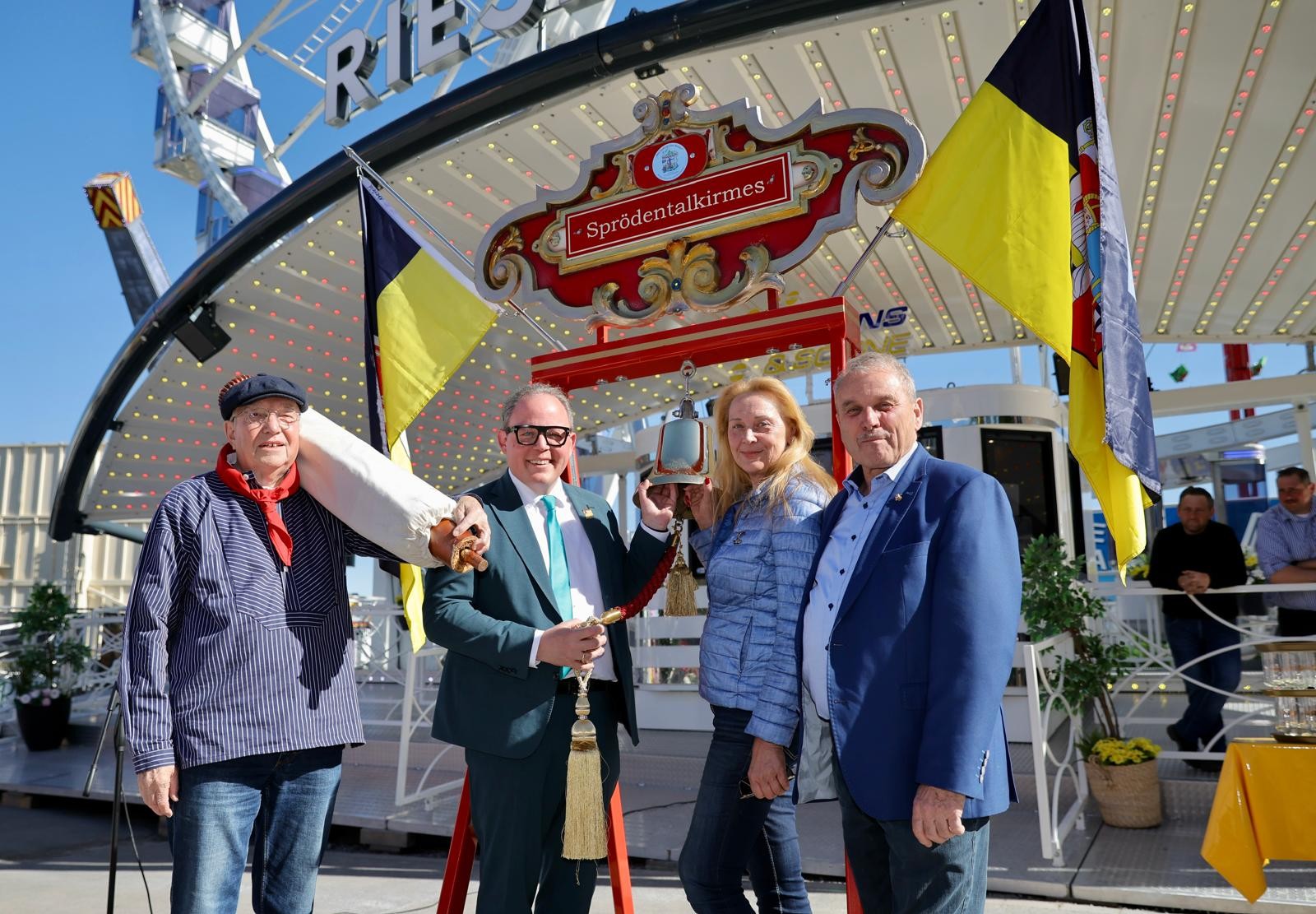 Vier Personen stehen vor dem Schild „Sprödentalkirmes“ und einer aufgehängten Glocke. Im Hintergrund sind ein Fahrgeschäft, Fahnen und ein Riesenrad zu sehen.