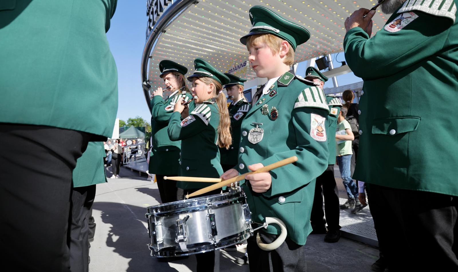 Kinder und Erwachsene in grünen Uniformen musizieren vor einem hell beleuchteten Fahrgeschäft. Im Vordergrund spielt ein Junge auf einer Trommel, weitere Kinder spielen Flöte.