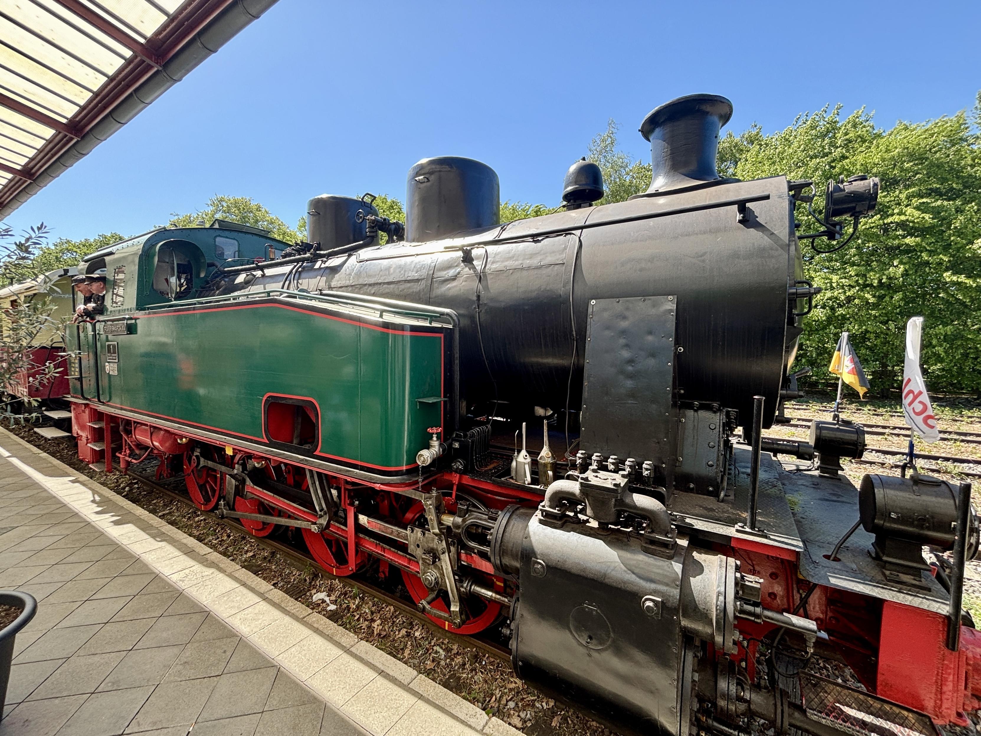 A vintage steam locomotive is displayed at a train station, featuring a green and black exterior with red wheels. A conductor is visible in the cab, and the background shows trees and a clear blue sky.