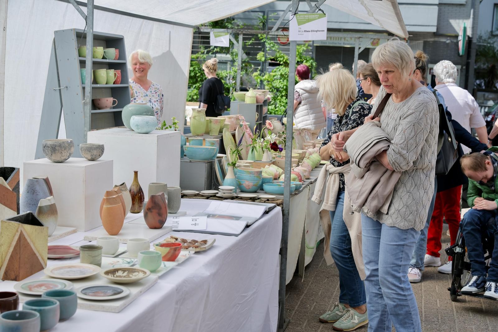 Ein Stand auf dem Pottbäckermarkt bei „Op dä Maat“ mit vielen Keramikstücken wie Schalen, Vasen, Tassen und Tellern. Mehrere Besucherinnen schauen sich die Auslage unter einem weißen Marktzelt an.
