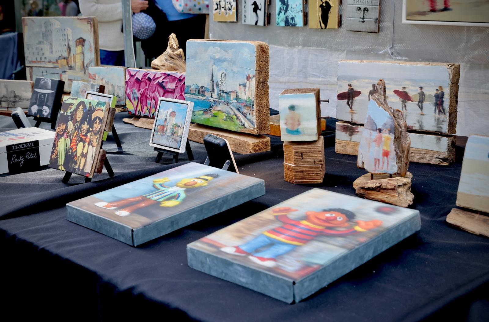 Auf einem Marktstand sind kleine Kunstwerke und bemalte Holzobjekte ausgestellt. Zu sehen sind unter anderem Stadtmotive, Figuren und farbenfrohe Bilder auf Holzblöcken.