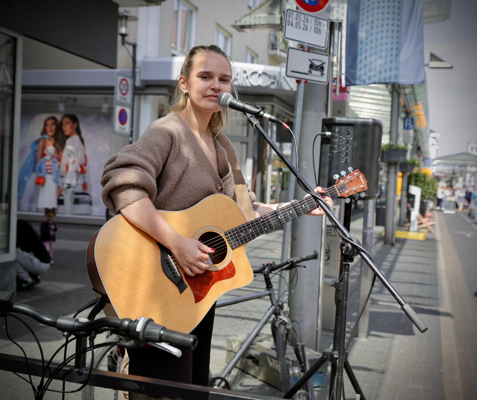Eine junge Musikerin steht mit Akustikgitarre an einem Mikrofon in der Innenstadt und spielt Straßenmusik. Im Hintergrund sind Geschäfte, Fahrräder und der Straßenraum zu sehen.