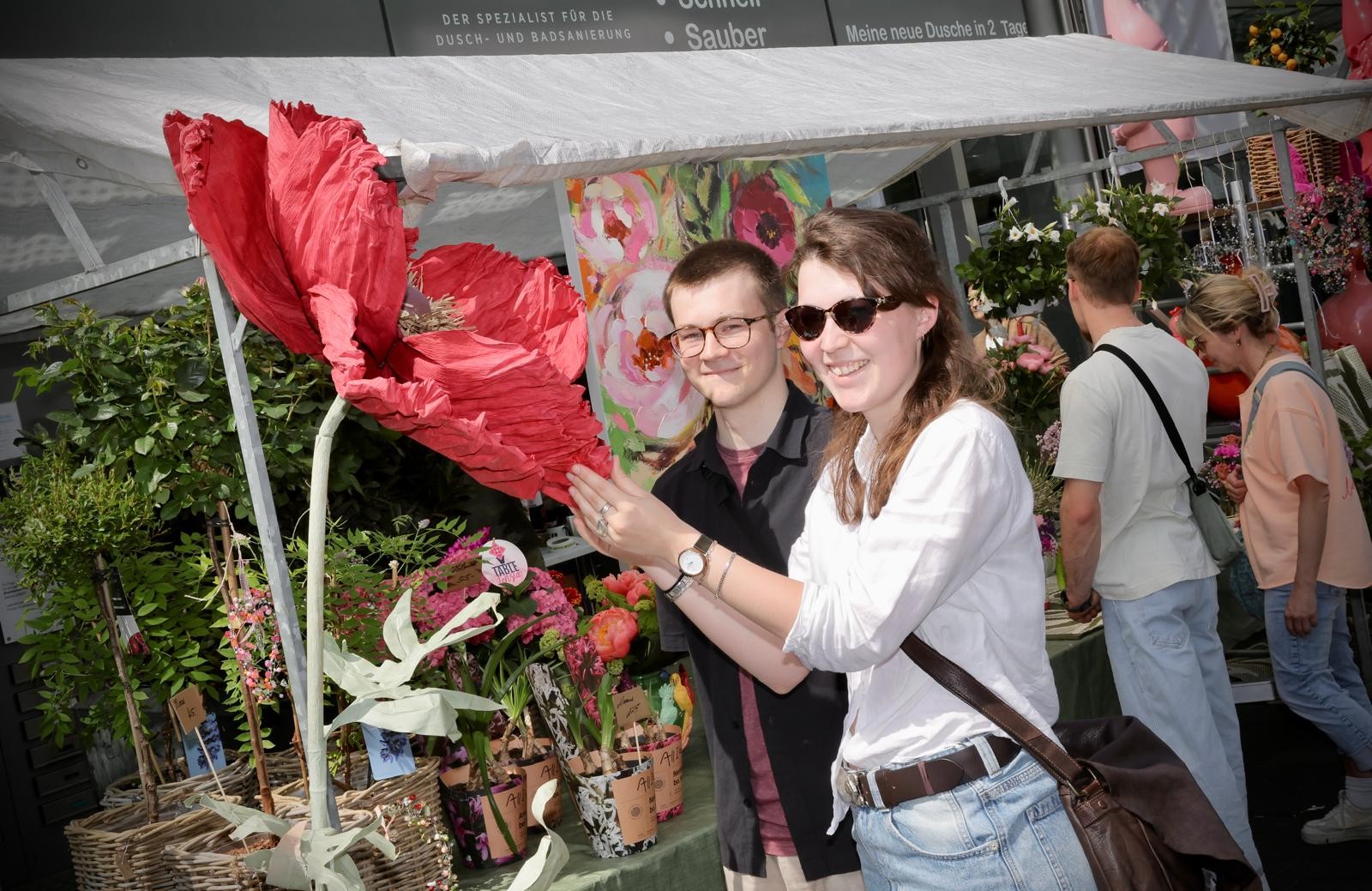 Ein Paar steht lachend an einem Blumenstand und hält eine große rote Papierblume in die Kamera. Um sie herum sind viele Pflanzen, Blumen und dekorative Artikel ausgestellt.