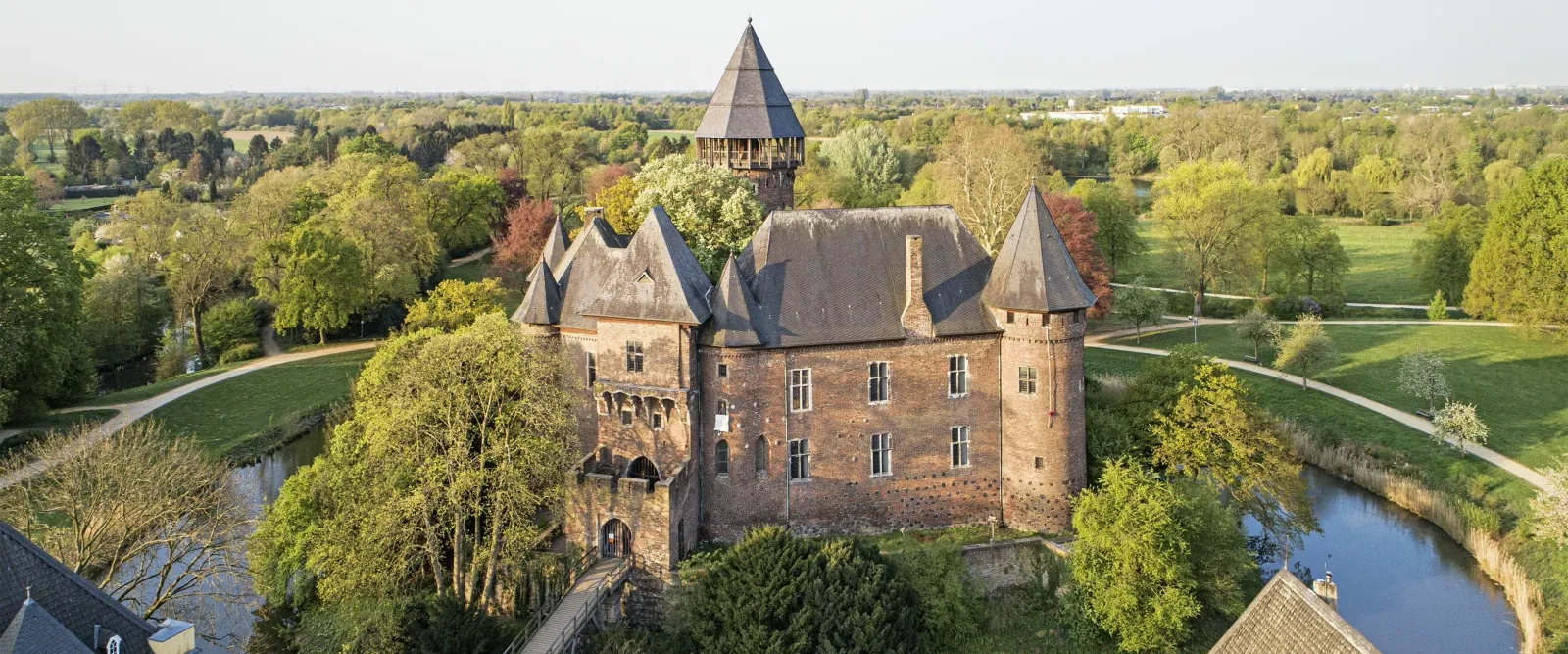 Luftaufnahme der Burg Linn in Krefeld. Die historische Backsteinburg mit mehreren Türmen ist von Wassergräben, Wegen und viel Grün umgeben. Im Hintergrund erstreckt sich eine weite Park- und Landschaftsfläche mit Bäumen.