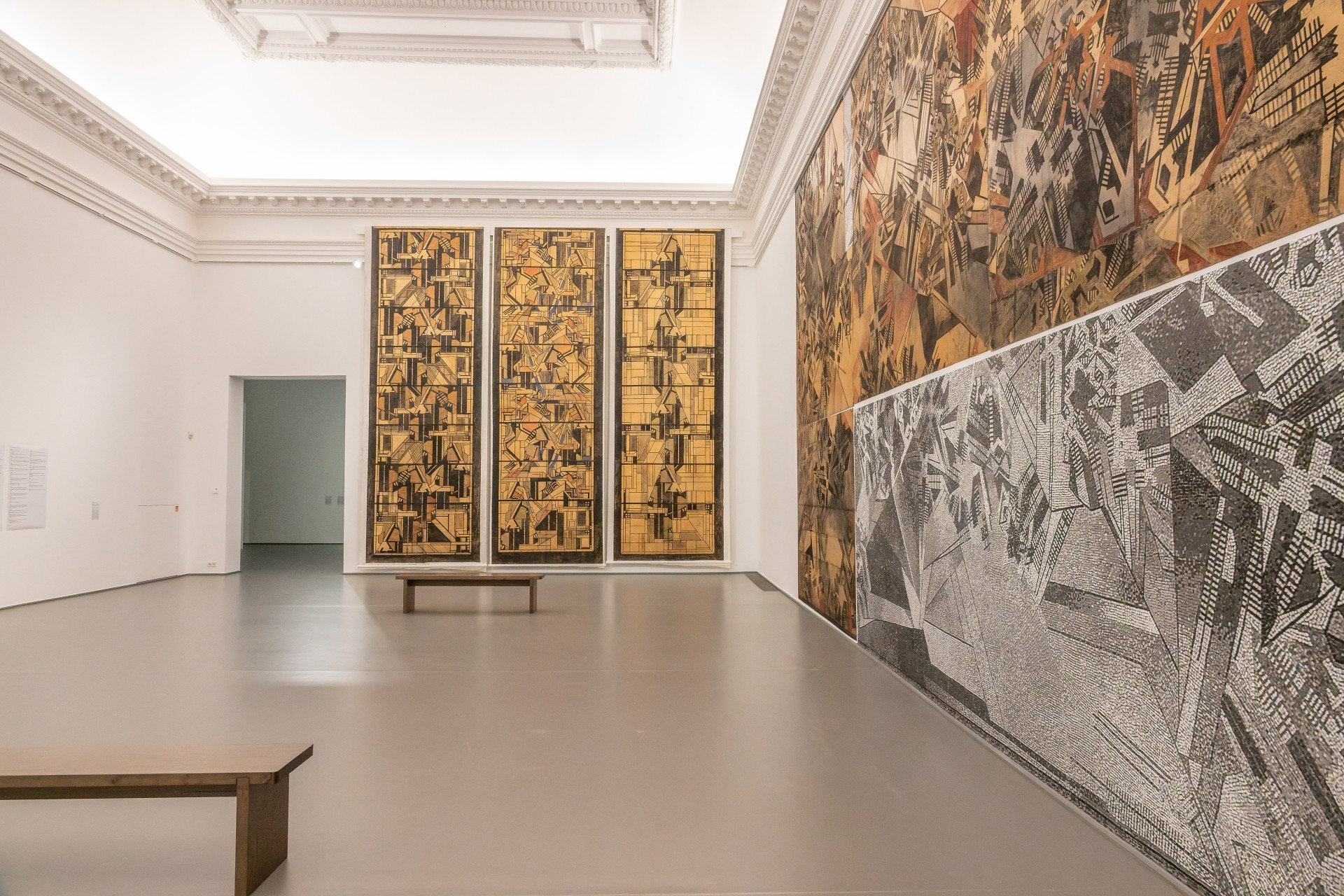 Heller Museumsraum mit großformatigen Wandarbeiten. Rechts ist ein langes, abstraktes Bild in Schwarz, Weiß und Brauntönen zu sehen, an der Stirnwand hängen drei hohe, geometrisch gestaltete Werke. In der Mitte steht eine schlichte Holzbank.