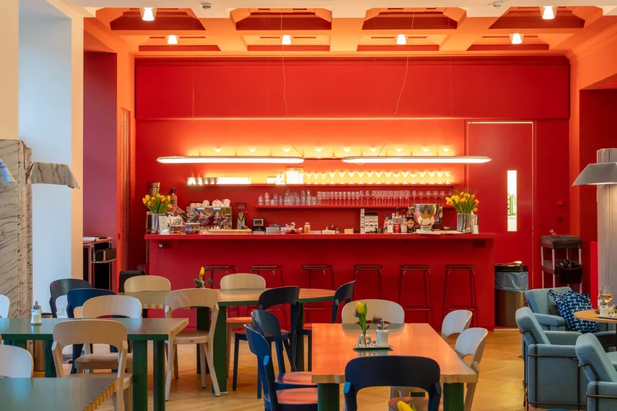 Cafébereich im Kaiser-Wilhelm-Museum mit roten Wänden, langer Theke, Tischen, Stühlen und Sofas. Warmes Licht und Blumen auf den Tischen sorgen für eine gemütliche Atmosphäre.
