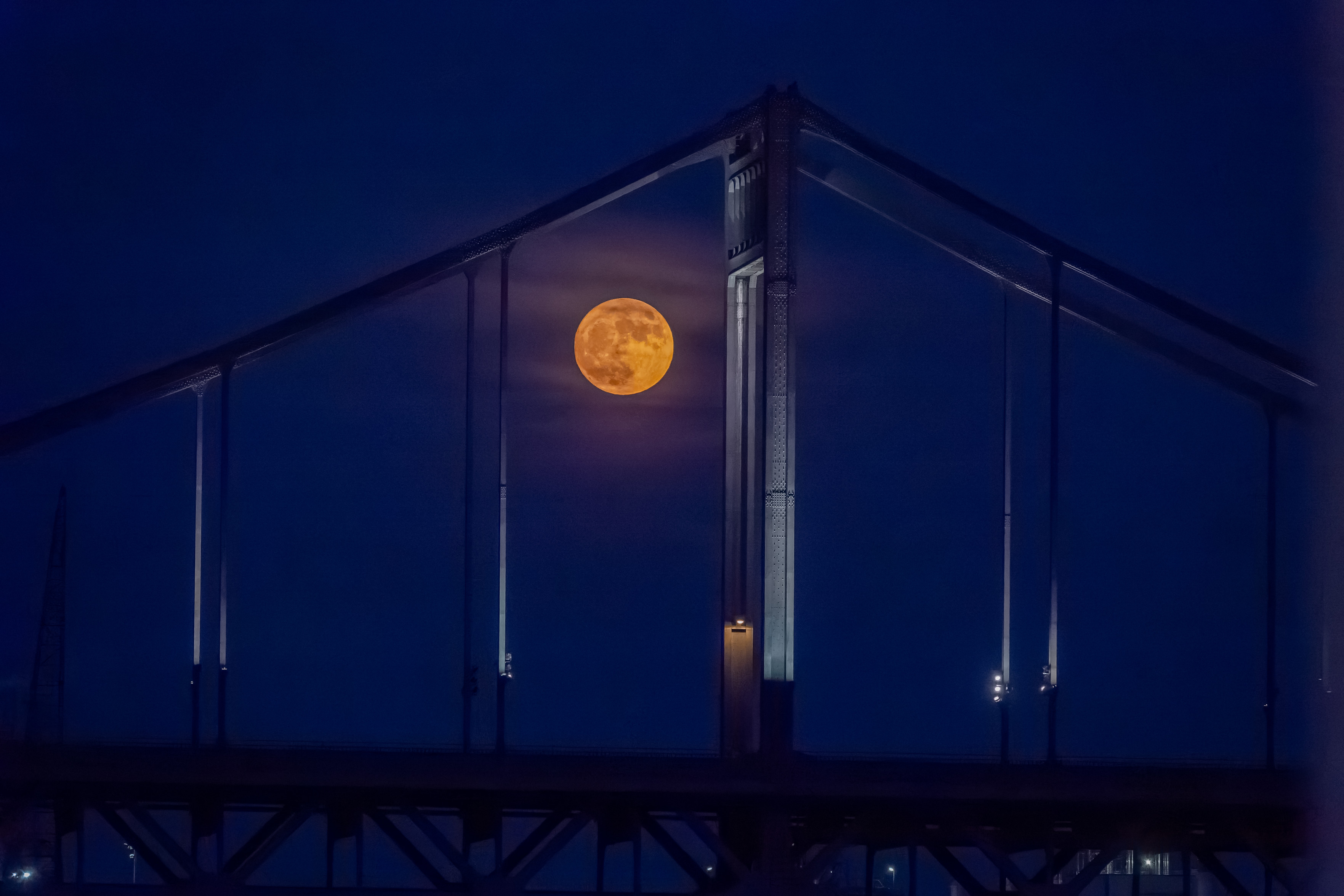 Nächtliche Aufnahme einer großen Brücke im blauen Dämmerlicht. Zwischen den Brückenseilen leuchtet ein orangefarbener Vollmond, während die Konstruktion als dunkle Silhouette zu sehen ist.