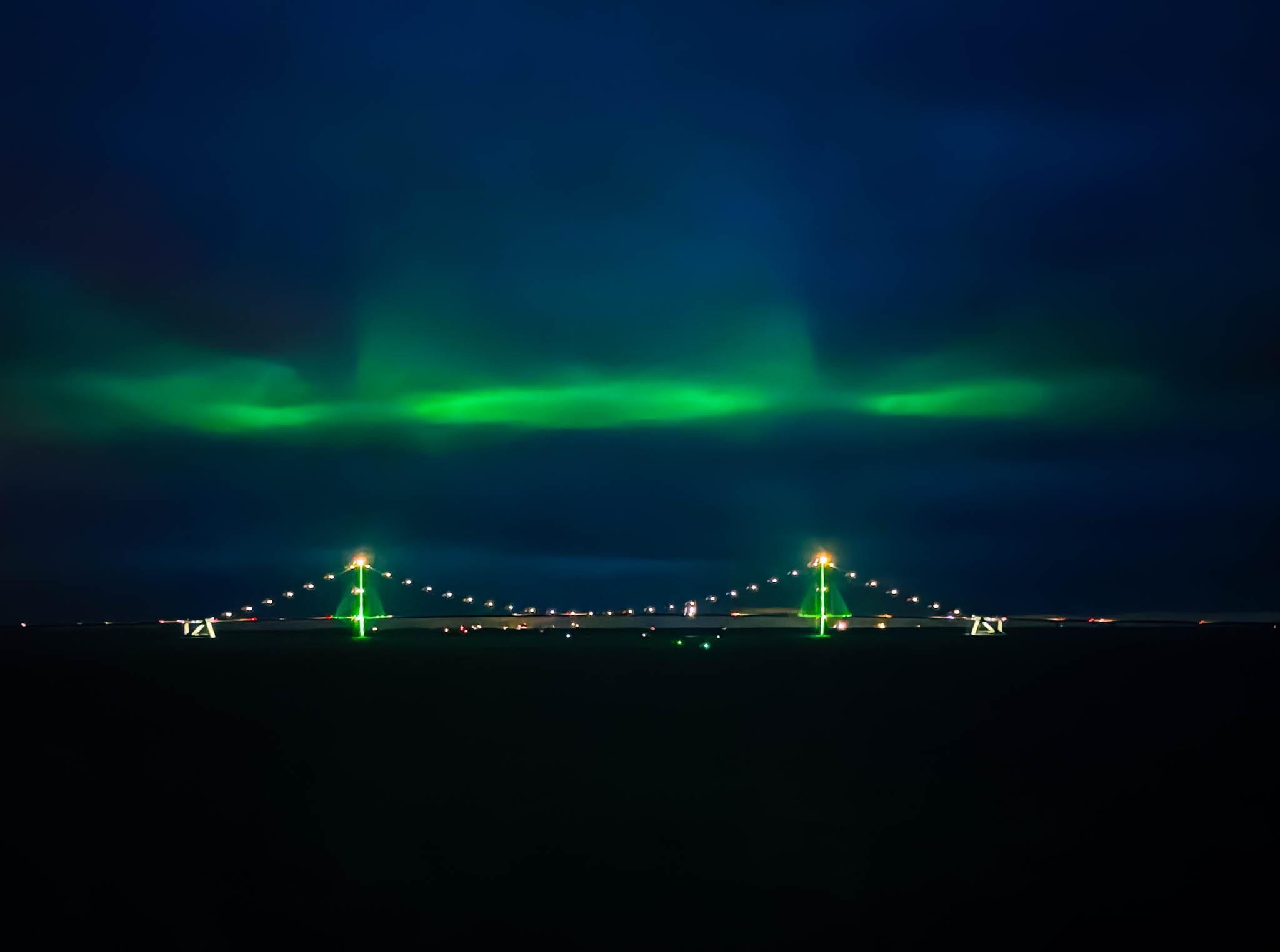 Eine Nachtszene mit einer Brücke, die mit grünen und gelben Lichtern beleuchtet wird, vor einem dunklen Himmel, der von leuchtenden grünen Polarlichtern erhellt wird.