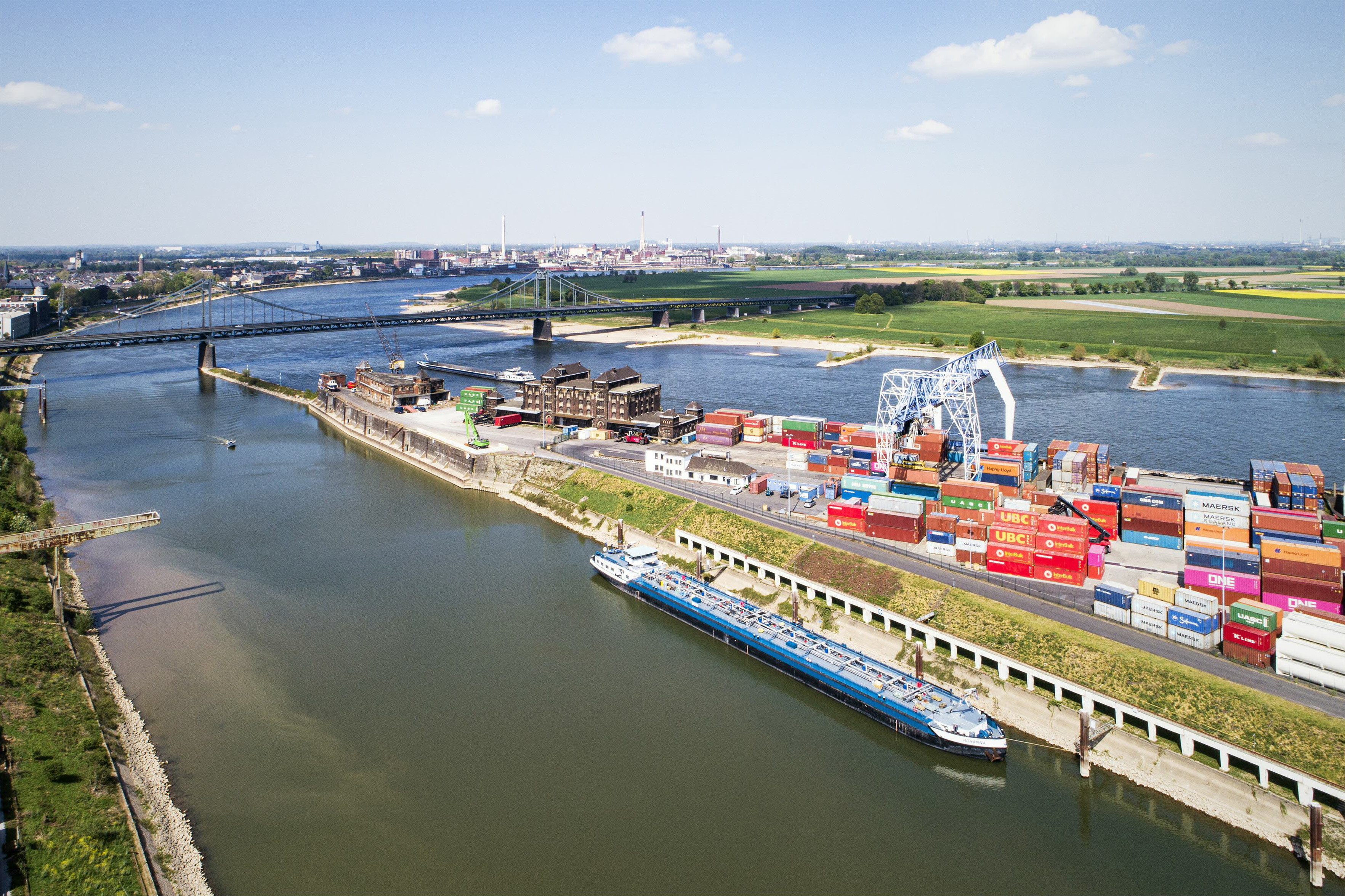Luftaufnahme des Krefelder Rheinhafens in Uerdingen mit Containerflächen, Hafenkran und einem Frachtschiff am Kai. Im Hintergrund sind der Rhein, die Rheinbrücke und weitläufige Felder unter blauem Himmel zu sehen.