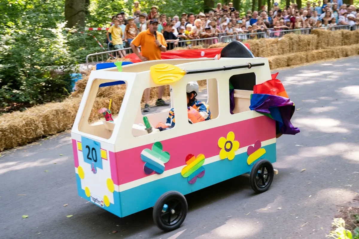 Eine bunt gestaltete Seifenkiste in Form eines kleinen Busses fährt auf einer Waldstrecke am Publikum vorbei. Die Strecke ist mit Strohballen abgesichert.
