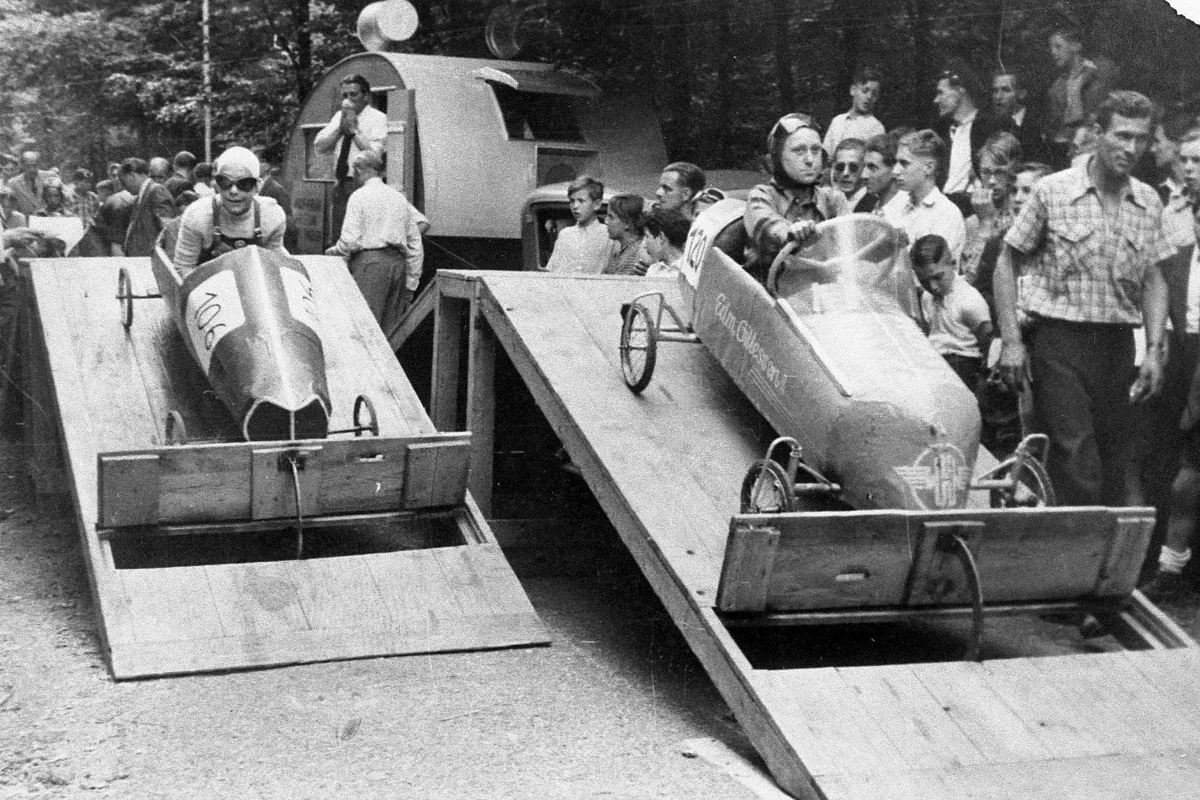 Historische Schwarz-Weiß-Aufnahme: Zwei Fahrer sitzen in Seifenkisten auf hölzernen Startrampen, während Zuschauerinnen und Zuschauer am Rand das Rennen verfolgen.