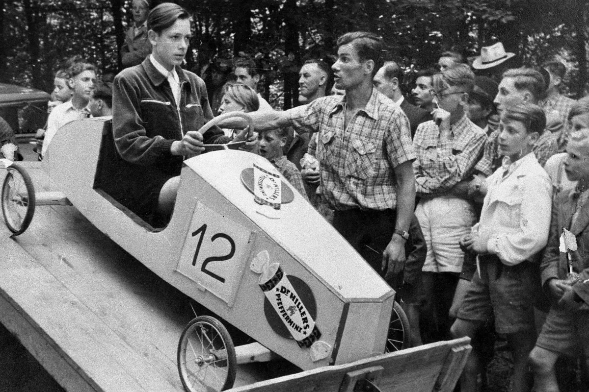 Historische Schwarz-Weiß-Aufnahme: Ein Junge sitzt in einer Seifenkiste mit der Startnummer 12, umringt von Kindern, Jugendlichen und Erwachsenen am Hülser Berg.