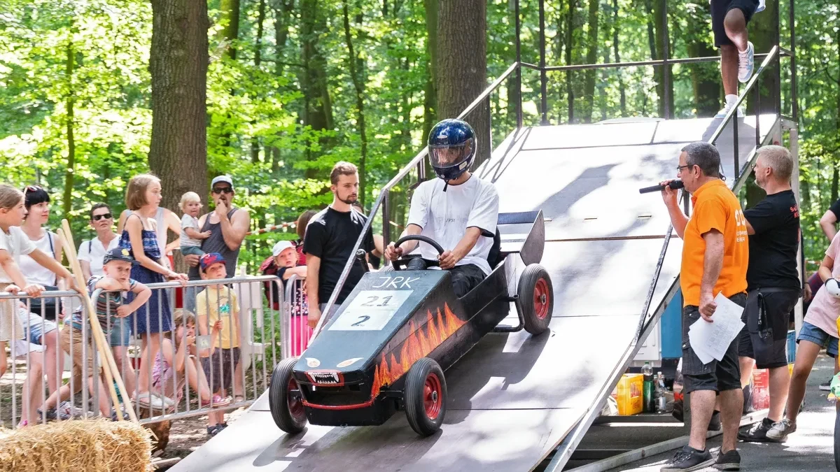 Ein Fahrer mit Helm sitzt in einer schwarzen Seifenkiste auf der Startrampe. Daneben stehen Helfer und Moderator, während Zuschauerinnen und Zuschauer am Rand der Waldstrecke warten.
