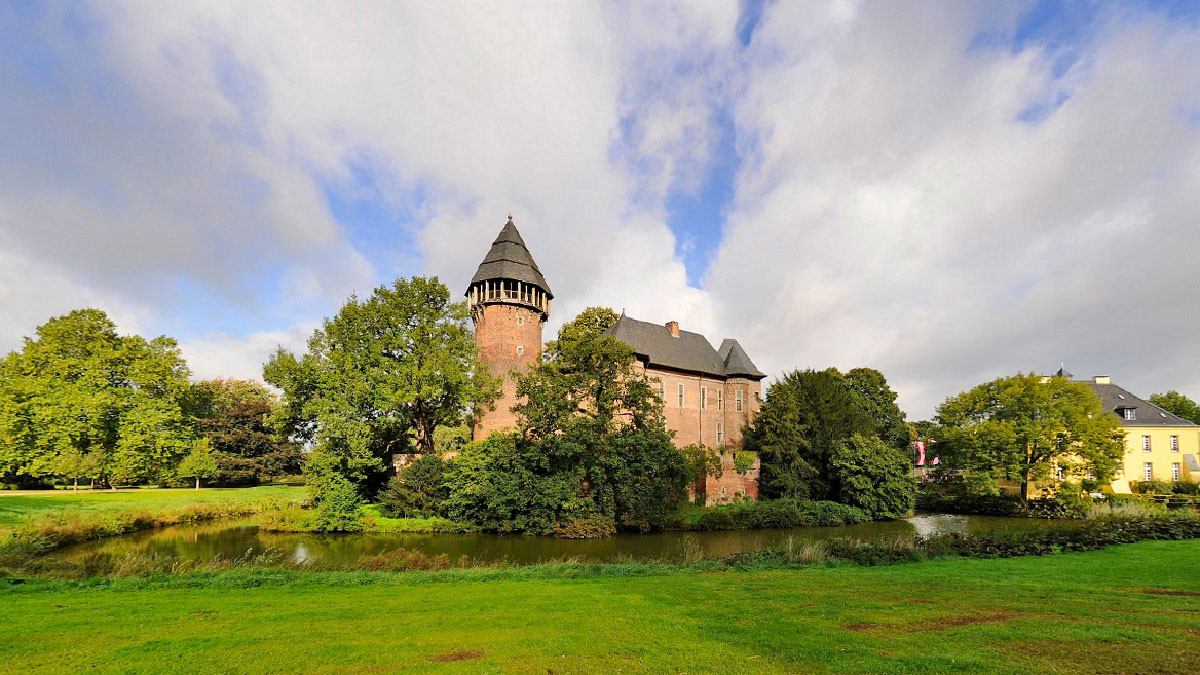 Ein historisches Backsteinschloss mit rundem Turm und spitzem Dach steht inmitten eines Parks, umgeben von alten Bäumen. Vor dem Schloss liegt ein Wassergraben, der die Gebäude und das Grün spiegelt. Rechts schließen sich weitere Gebäude mit hellen Fassaden an. Über der weitläufigen Rasenfläche spannt sich ein dramatischer Himmel mit großen Wolken und blauen Auflockerungen.