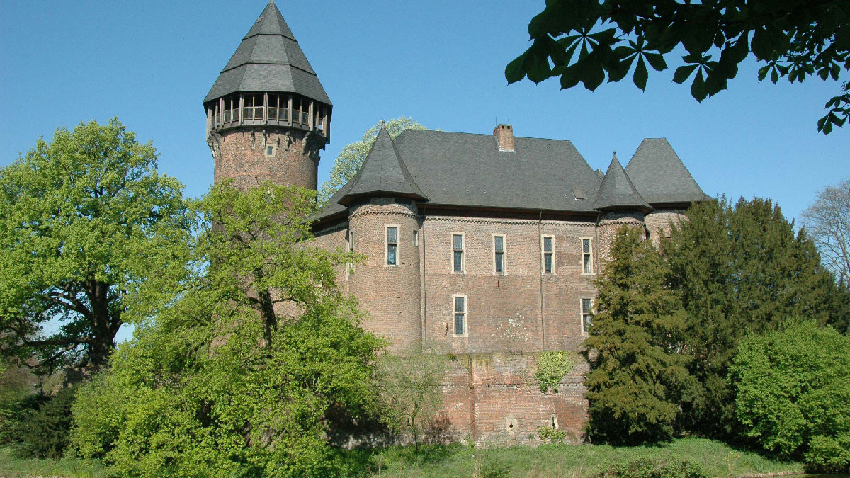 Eine Backsteinburg mit einem hohen Rundturm und einem steilen Dach steht an einem Wassergraben, umgeben von Bäumen unter einem klaren blauen Himmel.