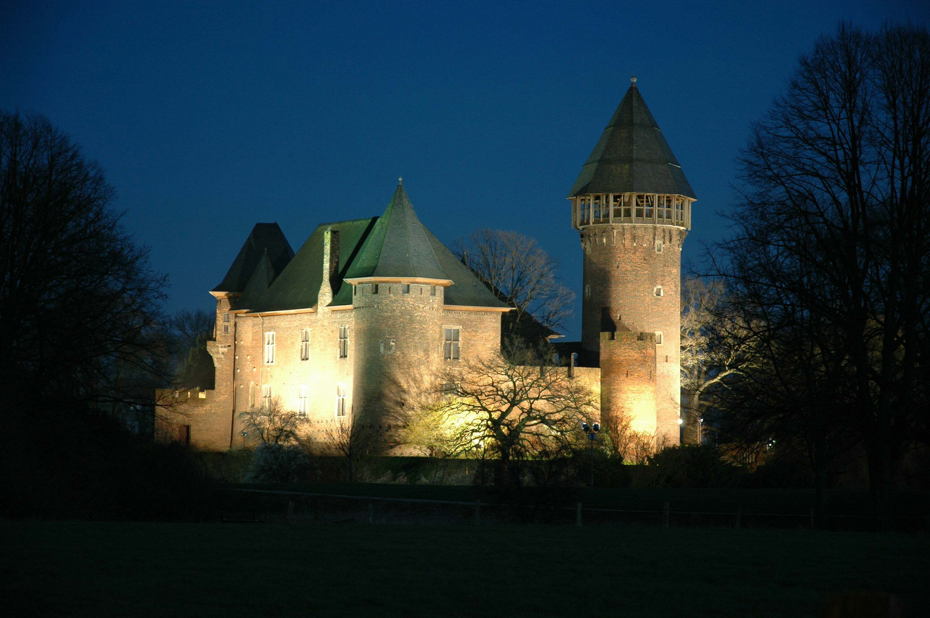 Eine große Backsteinburg mit einem hohen Rundturm wird nachts von Lichtern angestrahlt und ist von blattlosen Bäumen sowie einem dunklen Himmel umgeben.