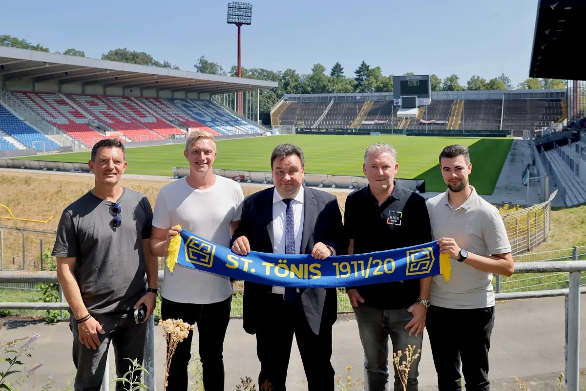 Fünf Männer stehen am Spielfeldrand der Grotenburg und halten gemeinsam einen blau-gelben Schal des SC St. Tönis 1911/20. Im Hintergrund sind der Rasen und die Tribünen des Stadions zu sehen.