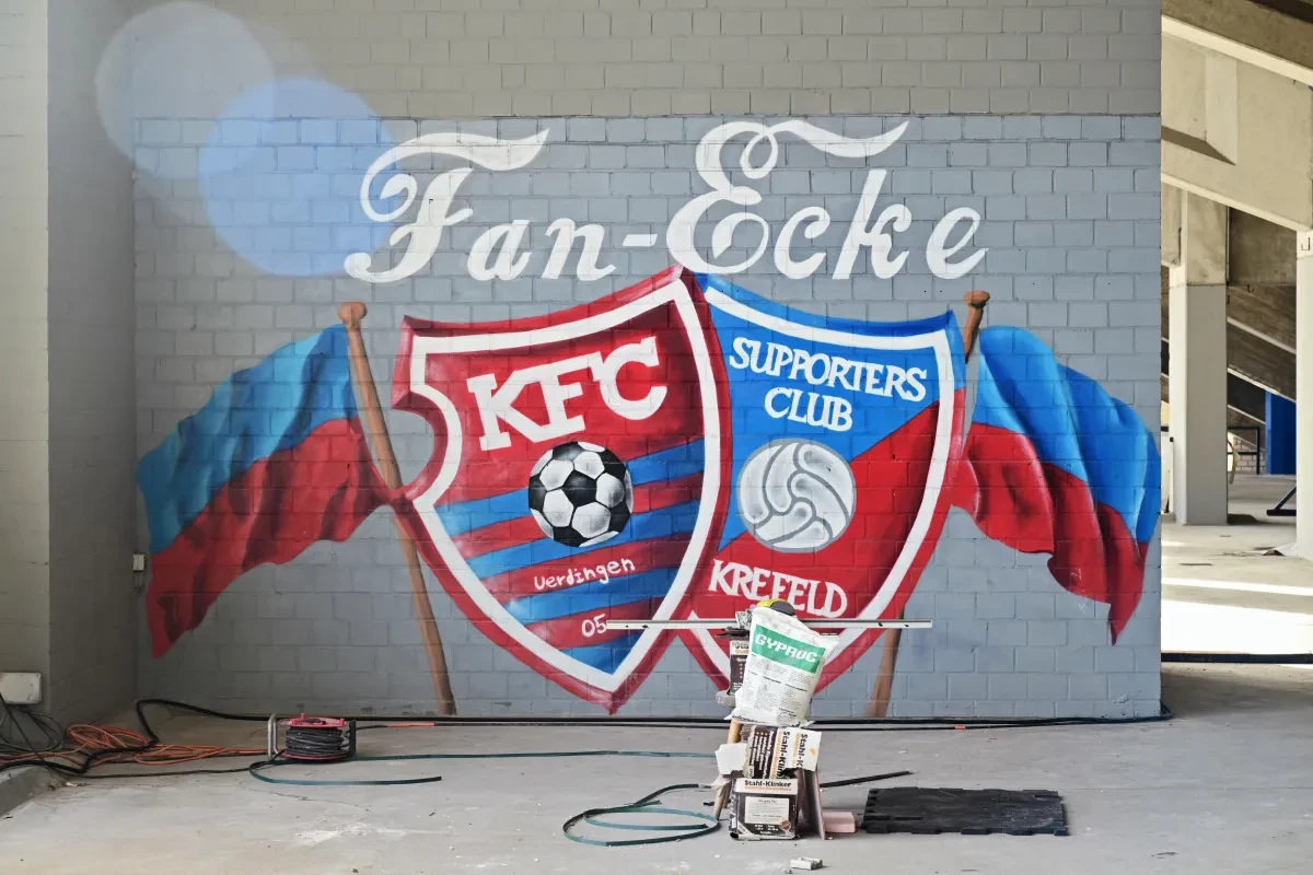 Wandgemälde in der Grotenburg mit der Aufschrift „Fan-Ecke“. Darunter sind die Wappen des KFC Uerdingen 05 und des Supporters Club Krefeld vor rot-blauen Fahnen dargestellt.