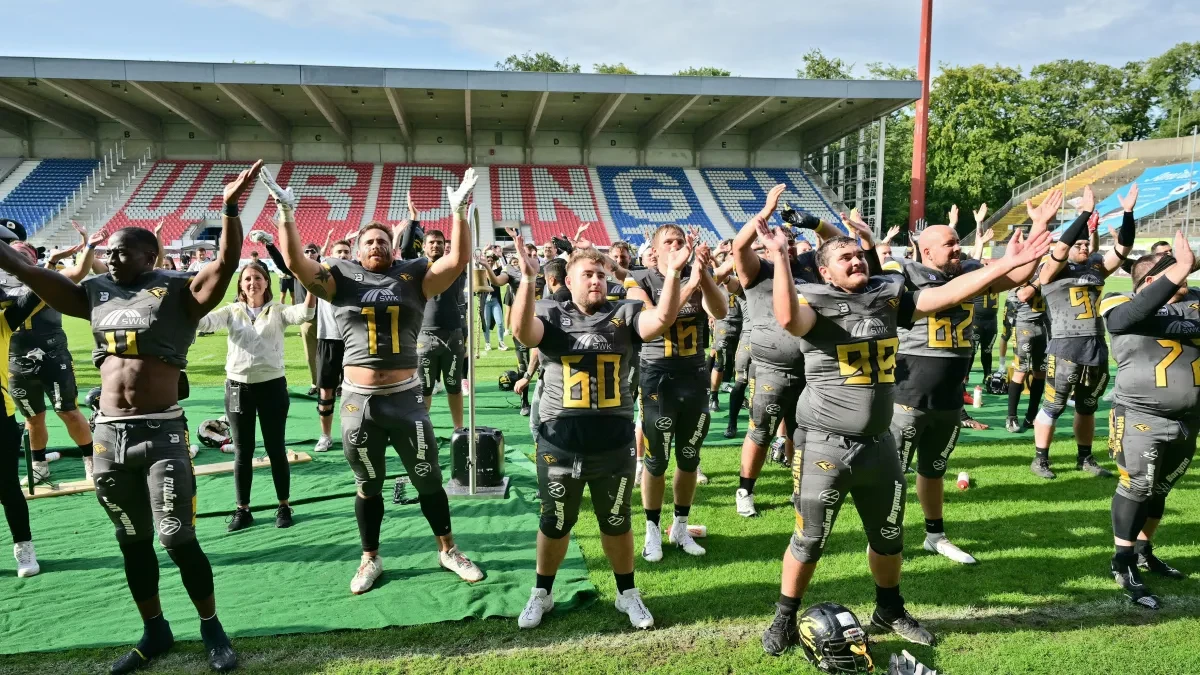 Spieler eines American-Football-Teams in schwarzen Trikots stehen nach einem Spiel auf dem Rasen der Grotenburg und applaudieren mit erhobenen Armen. Auf der Tribüne im Hintergrund ist der Schriftzug „Uerdingen“ zu sehen.