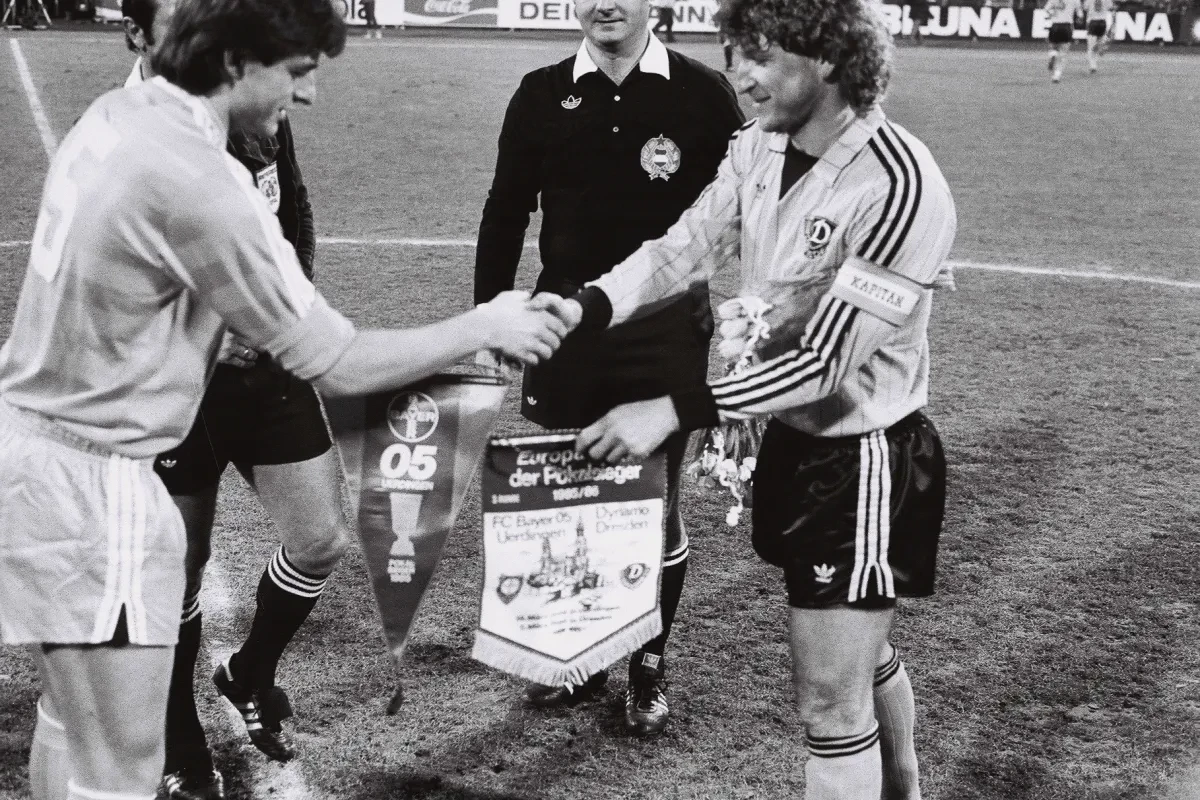Historische Schwarz-Weiß-Aufnahme vor einem Fußballspiel: Zwei Mannschaftskapitäne reichen sich auf dem Spielfeld die Hand und tauschen Wimpel aus. Zwischen ihnen stehen die Schiedsrichter.