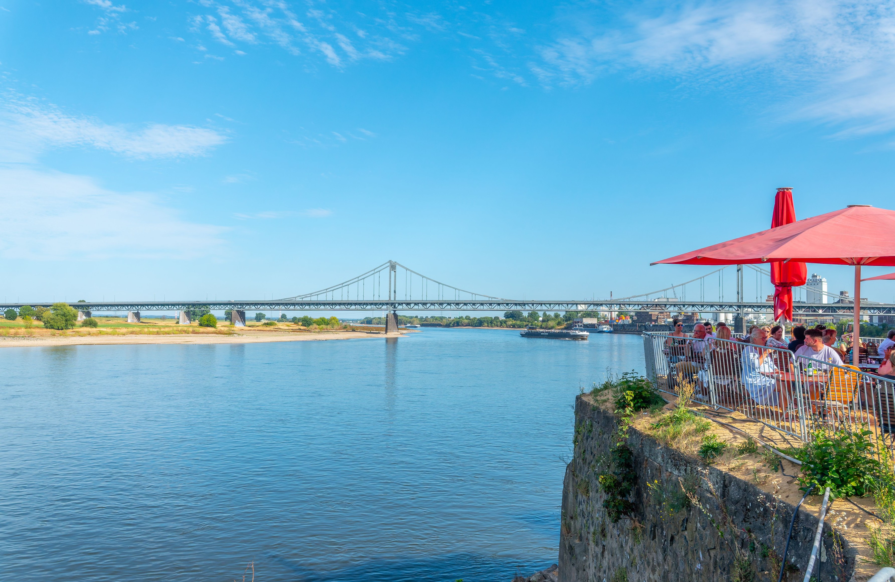 Blick über den Rhein auf die Uerdinger Rheinbrücke unter blauem, leicht bewölktem Himmel. Rechts sitzen zahlreiche Menschen auf einer Außenterrasse unter großen roten Sonnenschirmen direkt am Flussufer. Auf dem Wasser fährt ein Schiff.