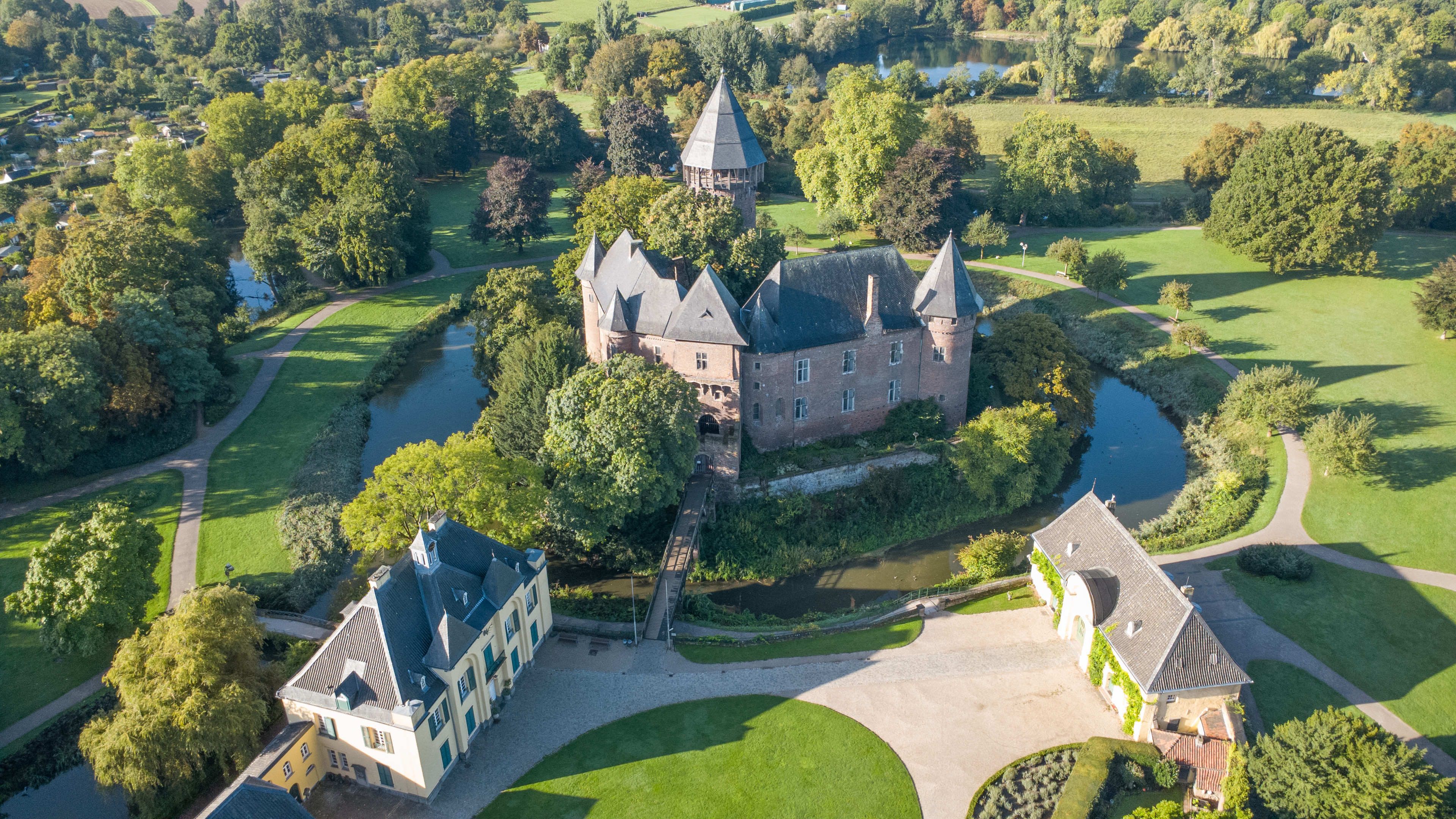 Luftaufnahme der Burg Linn inmitten einer weitläufigen, grünen Parkanlage. Die historische Wasserburg ist von einem Burggraben umgeben und über eine Brücke mit der Vorburg verbunden. Im Hintergrund sind weitere Wege, Bäume und Wasserflächen zu sehen.