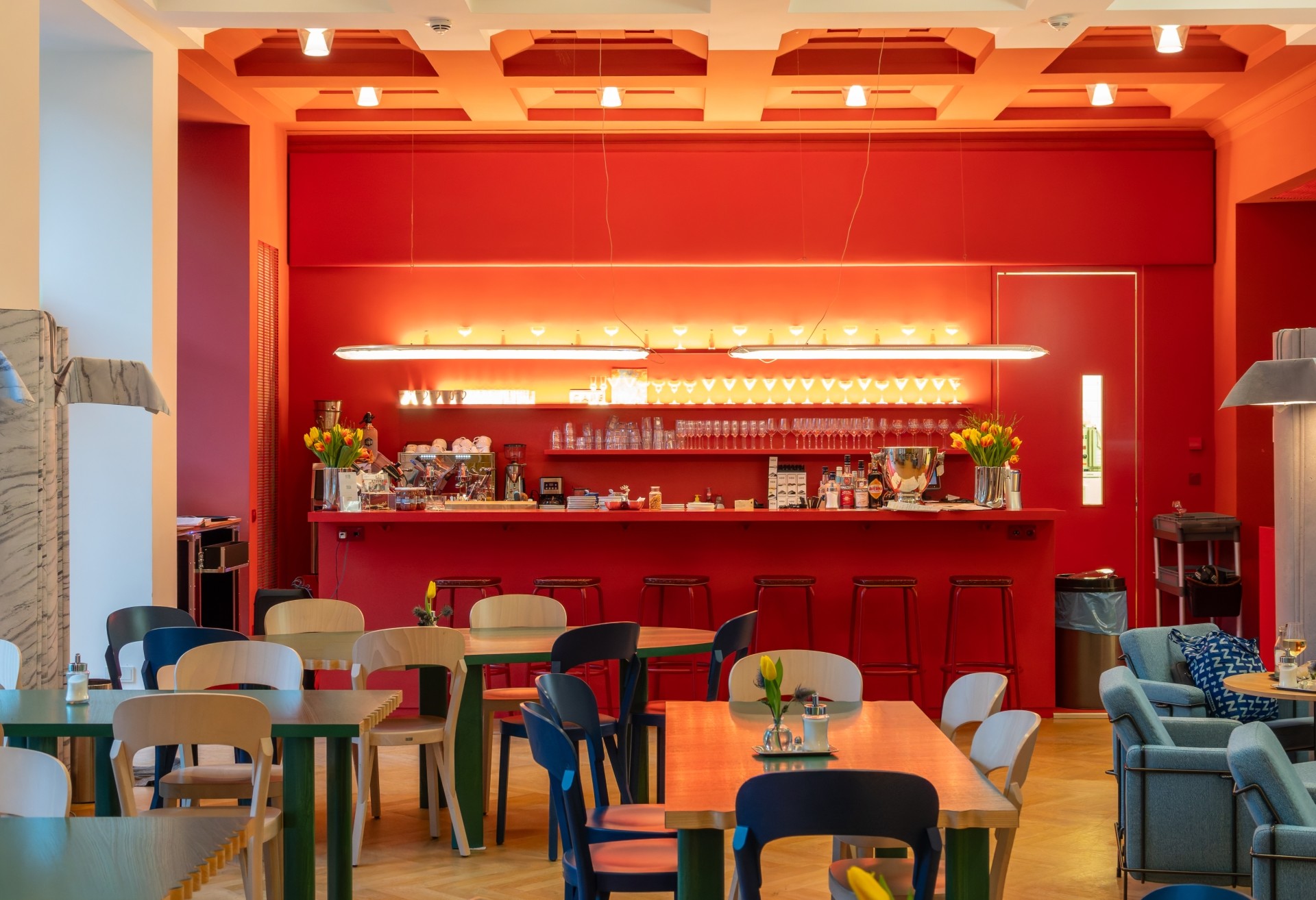 Cafébereich des Kaiser-Wilhelm-Museums mit leuchtend roter Bar, beleuchteten Regalen, Tischen, Stühlen und gemütlichen Sesseln. Kleine Blumenvasen stehen auf den Tischen.