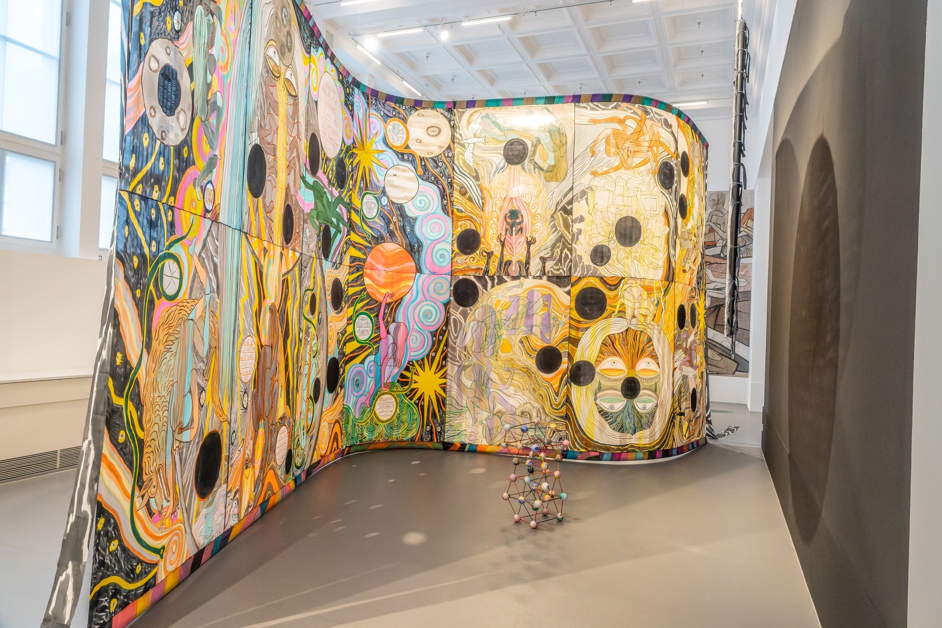 Großflächige, farbenfrohe Kunstinstallation in einem Museumssaal. Die geschwungenen Wände sind mit abstrakten Figuren, Mustern, Kreisen und leuchtenden Farben bemalt. Davor steht eine kleine filigrane Skulptur.