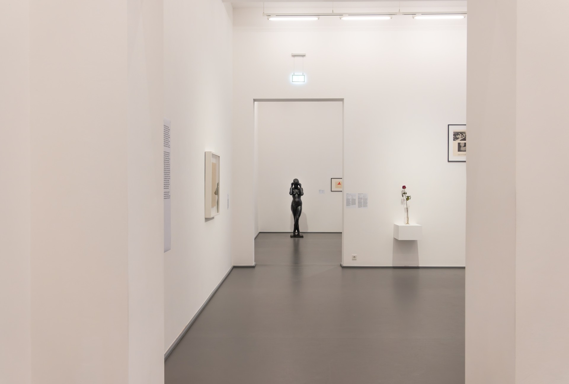 Blick durch mehrere Durchgänge in einen hellen, weiß gestalteten Ausstellungsraum des Kaiser-Wilhelm-Museums. An den Wänden hängen einzelne Kunstwerke und Informationstexte. Im hinteren Raum steht zentral eine dunkle menschliche Skulptur, rechts ist eine einzelne rote Rose in einer Vase ausgestellt.