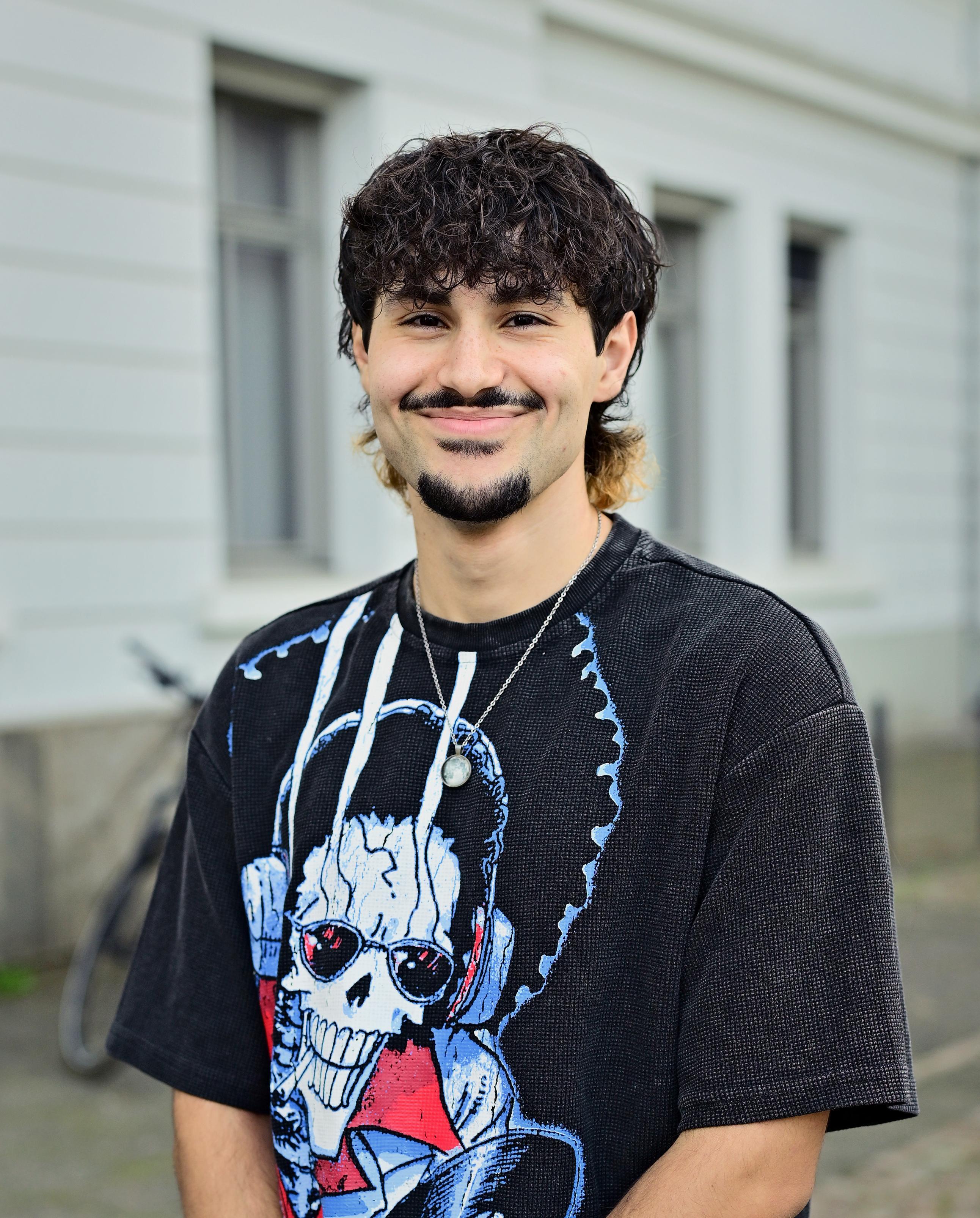 Ein junger Mann mit dunklen, lockigen Haaren, Schnurrbart und Kinnbart lächelt in die Kamera. Er trägt ein schwarzes T-Shirt mit einem großflächigen, bunten Totenkopf-Motiv sowie eine silberfarbene Halskette mit rundem Anhänger. Im unscharfen Hintergrund ist die helle Fassade eines Gebäudes mit mehreren Fenstern zu sehen.