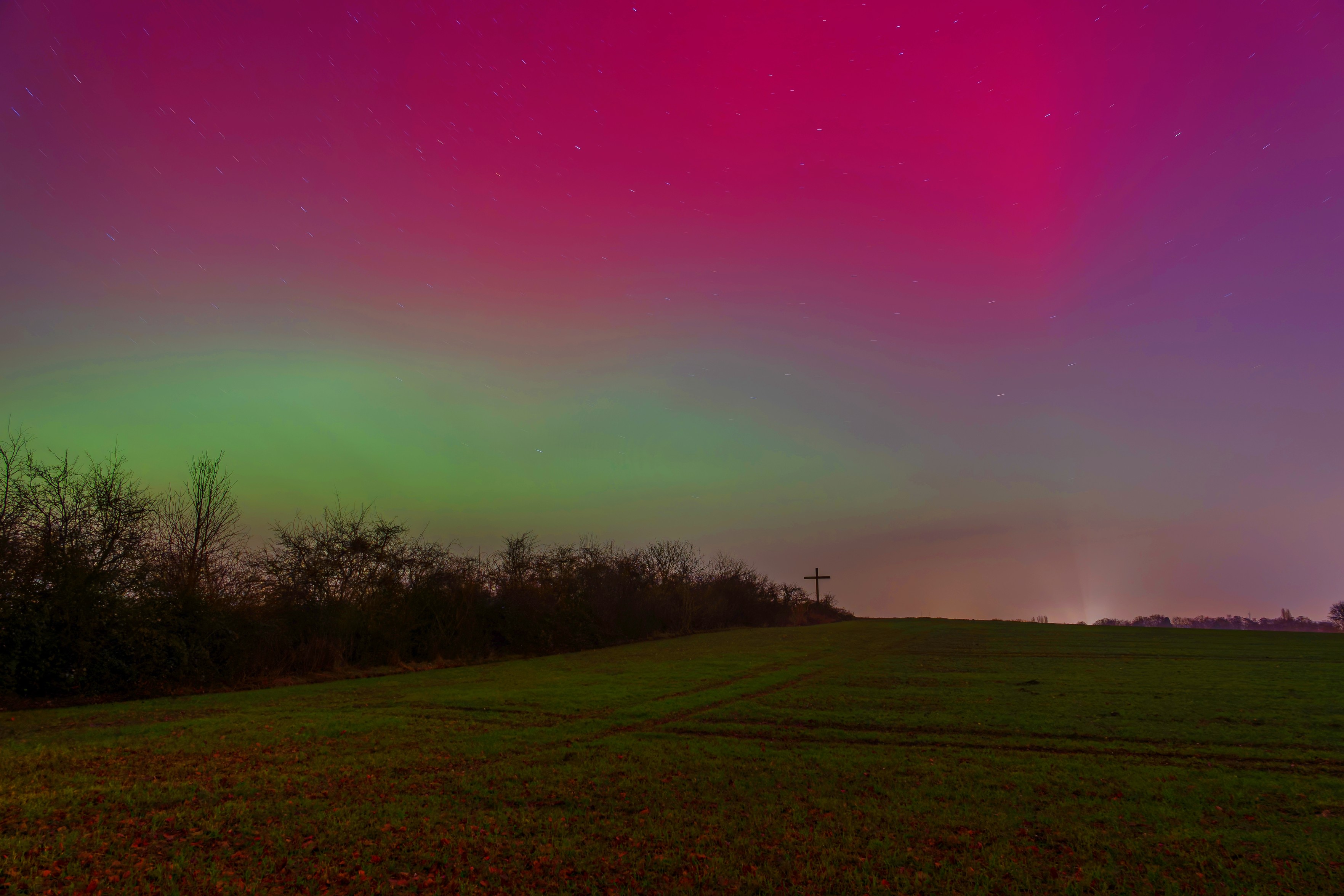 Nachthimmel über einem dunklen Feld: Ein breites Polarlichtband leuchtet grün, darüber färbt sich der Himmel pink-violett; Sterne sind als kurze Lichtspuren zu sehen. Am Horizont zeichnen sich eine Baumreihe und ein kleines Kreuz auf einer Anhöhe als Silhouette ab.