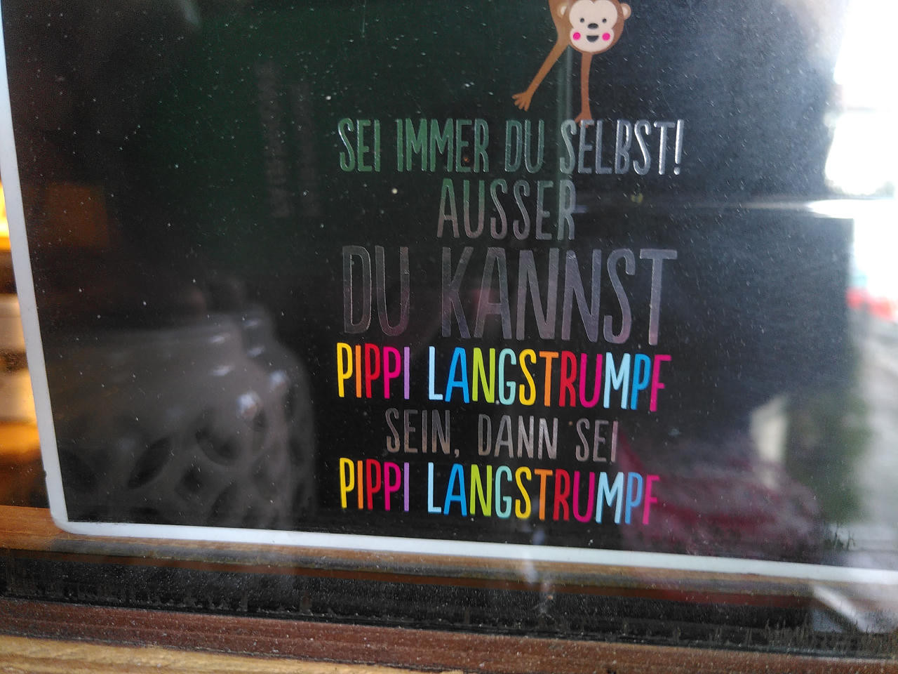 Hinter einer teilweise spiegelnden Glasscheibe steht eine Postkarte mit schwarzem Hintergrund. Der Text dieses Posts steht auf der Karte. Die Worte 