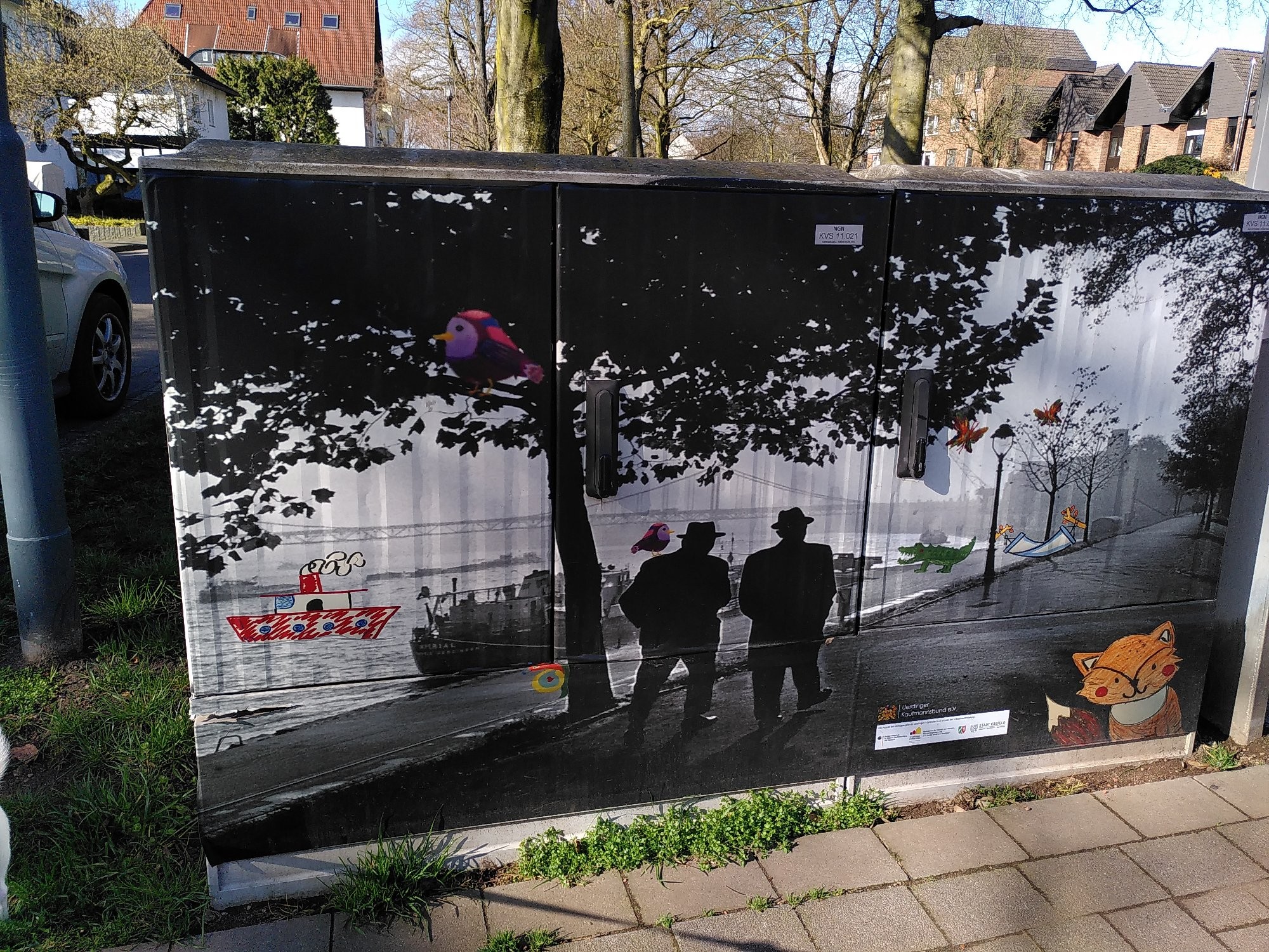 Ein Stromverteilerkasten, der mit einem Blid bemalt ist. Das Bild zeigt eine fotorealistische Szene am Ufer mit zwei Personen unter einem Baum in schwarzweiss. In bunten Farben sind Details hineingezeichnet, wie von Kinderhand. Ein Boot auf dem Wasser, ein Vogel im Baum, ein Wurm am Baumstamm, ein Krokodil auf der Ufermauer