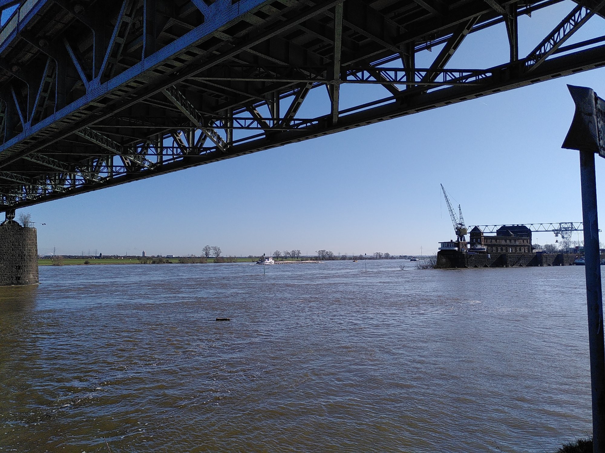 Unter einer Stahlbrücke hindurch sehen wir aufs Wasser. Im Hintergrund rechts eine Hafenanlage mit einem Kran.