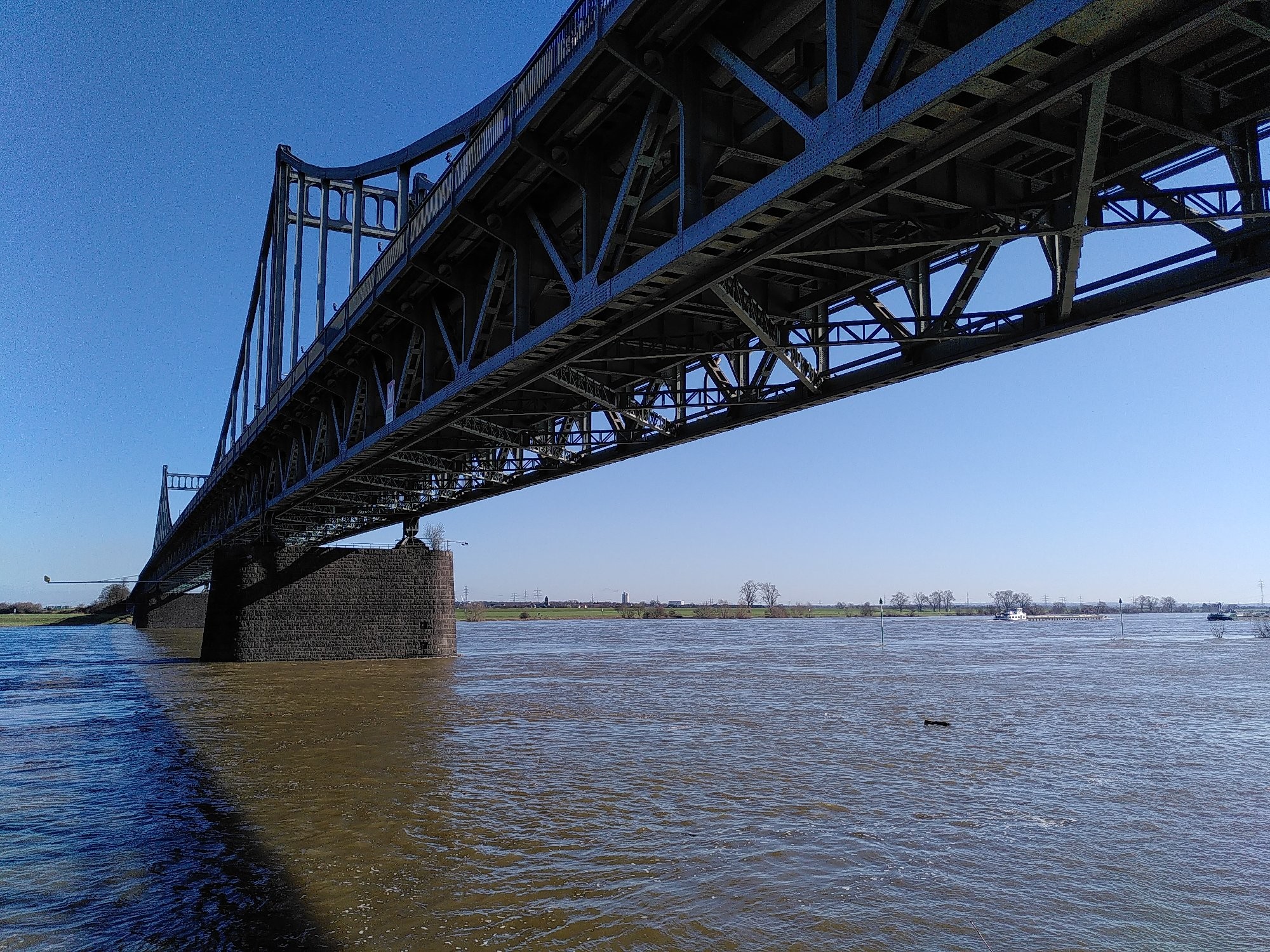 Über einer Wasserfläche und vor blauem Himmel sehen wir von unten die gewaltige Stahlkonstruktion einen Brücke