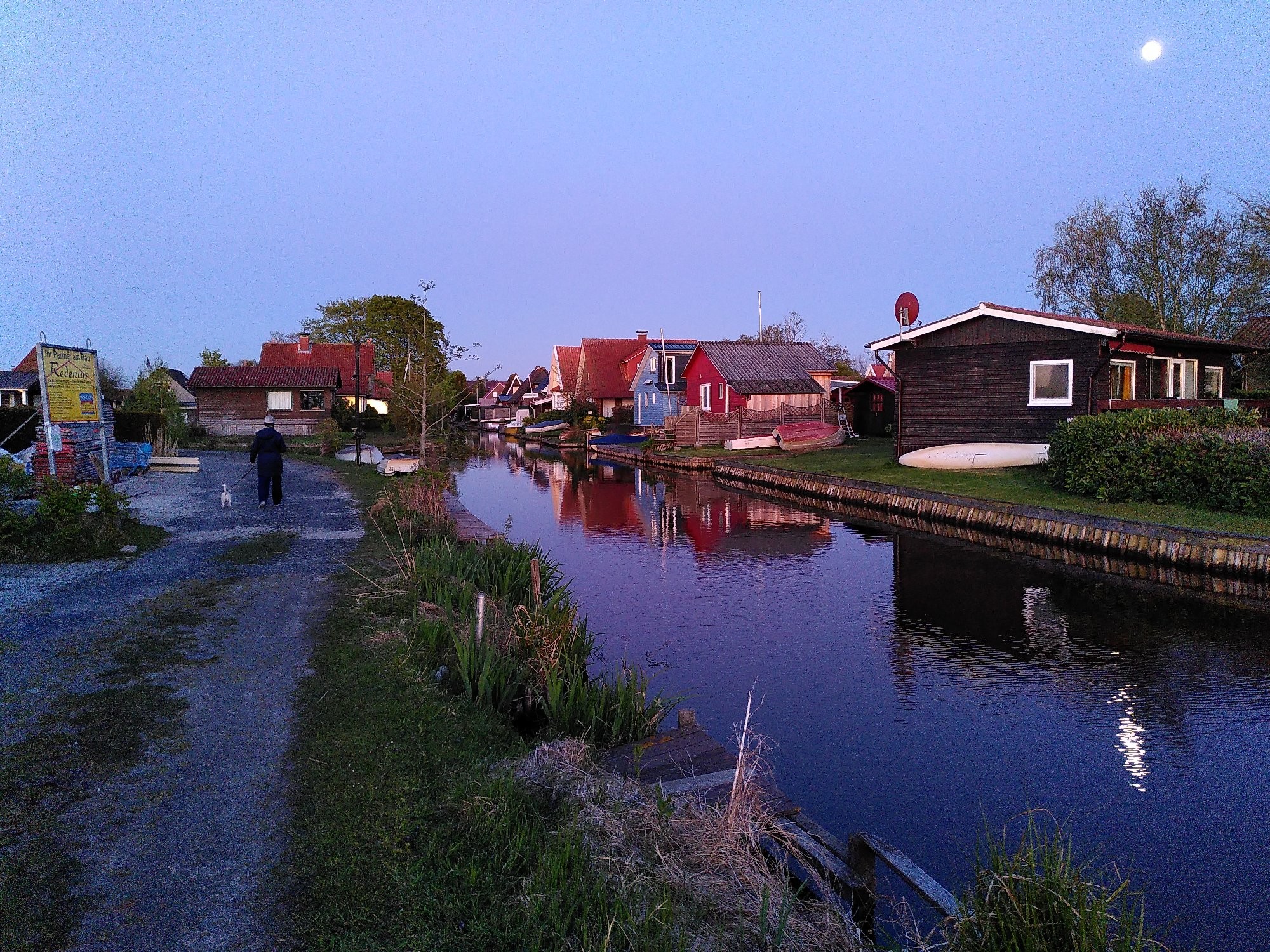 Ein Kanel führt rechts am Standort vorbei. Auf der anderen Seite sieht man bunte Holzhäuser in den Farben braun, rot und blau. Am Ufer liegen Boote. Abendstimmung. 