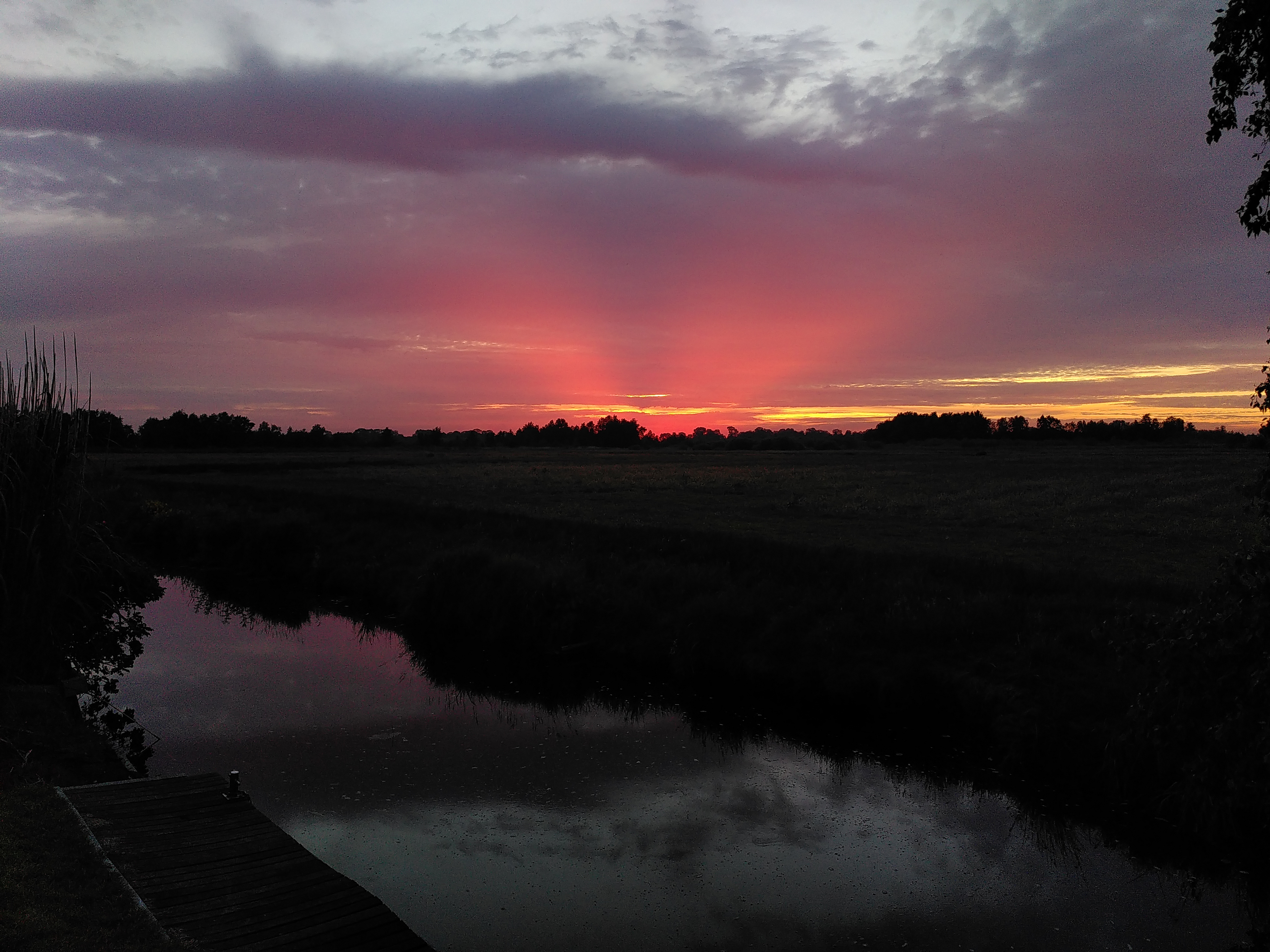 Sonnenuntergang über einem Kanal. Oben dunkle Wolken, das Orange des Sonnenuntergangs strahlt im die Wolken aus.