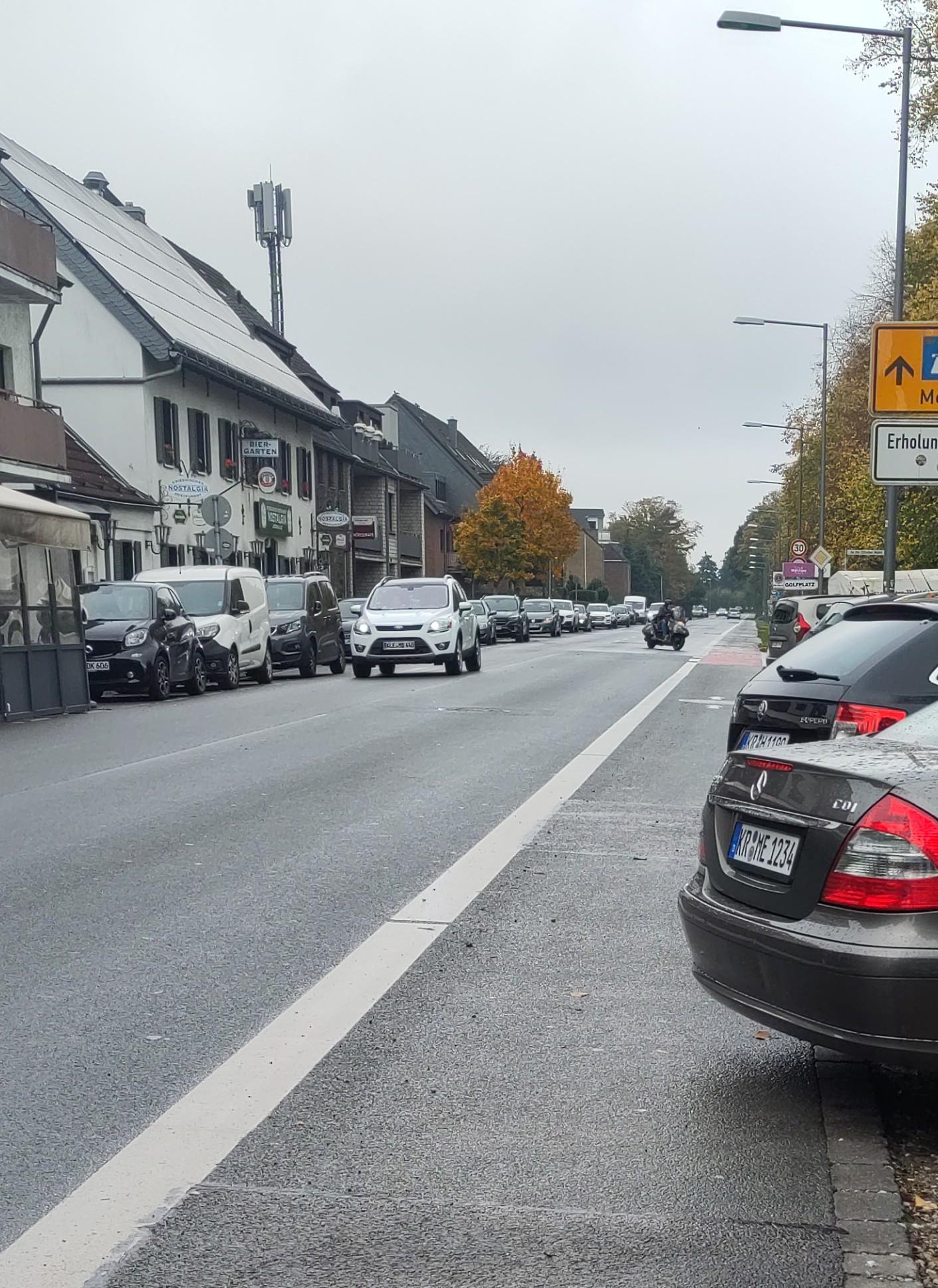 Blick entlang eines Fahrrad/Fußwegstreifens am rechten Straßenrand. Am linken Straßenrand mehrere Gebäude und parallel geparkte Autos. Rechts ragen mehrere Fahrzeuge, u.a. ein grauer Mercedes und ein schwarzer Skoda, mit dem Heck deutlich in den Fahrradstreifen hinein.