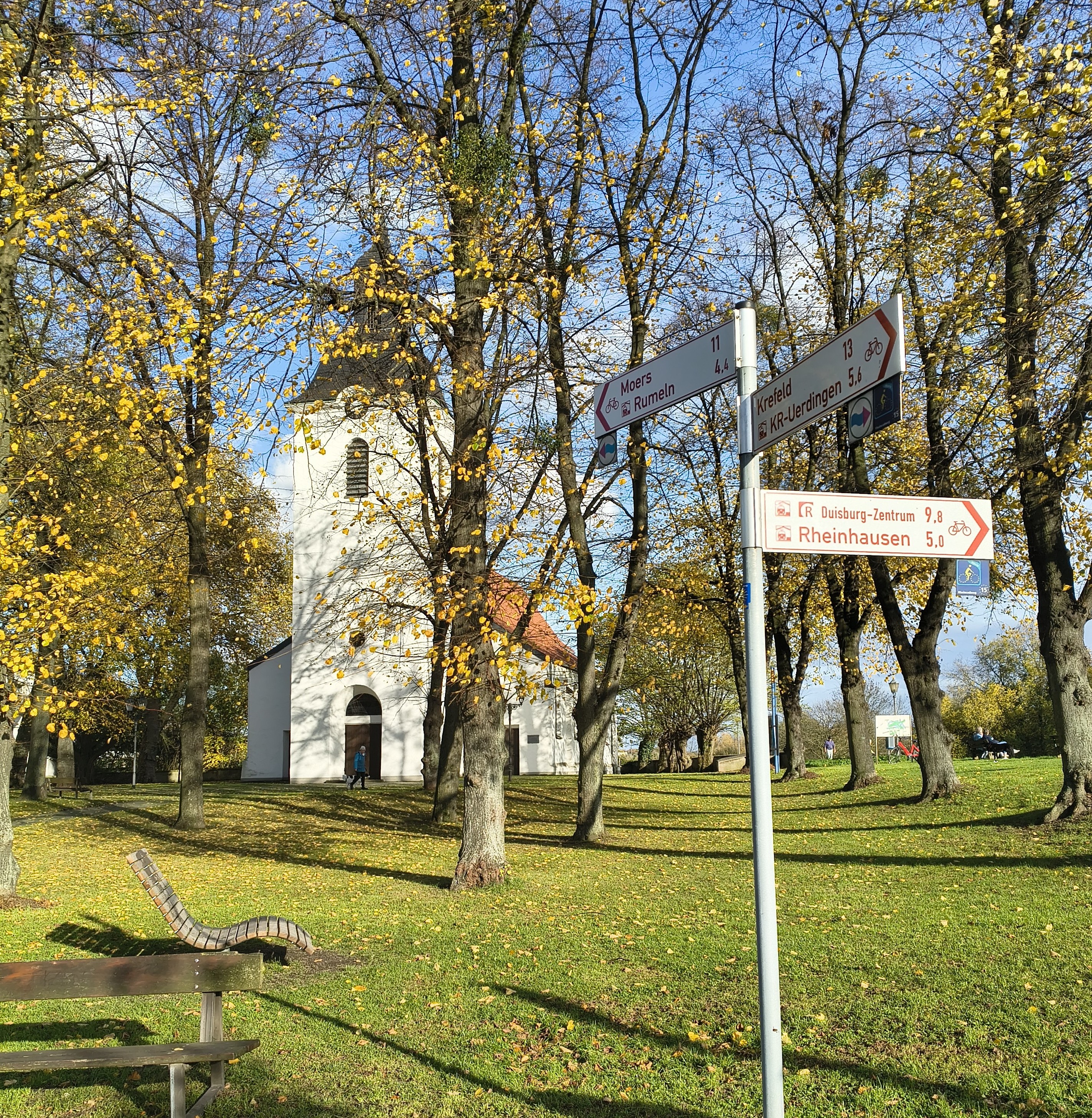 Weitläufige Wiese vor einer weißen, historischen Kirche mit rotem Dach. Die Bäume haben leuchtendes gelbes und grünes Herbstlaub. Im rechten Vordergrund steht ein brauner Wegweiser mit roten Fahrradsymbolen, und links eine hölzerne Sonnenliege. Blauer Himmel.