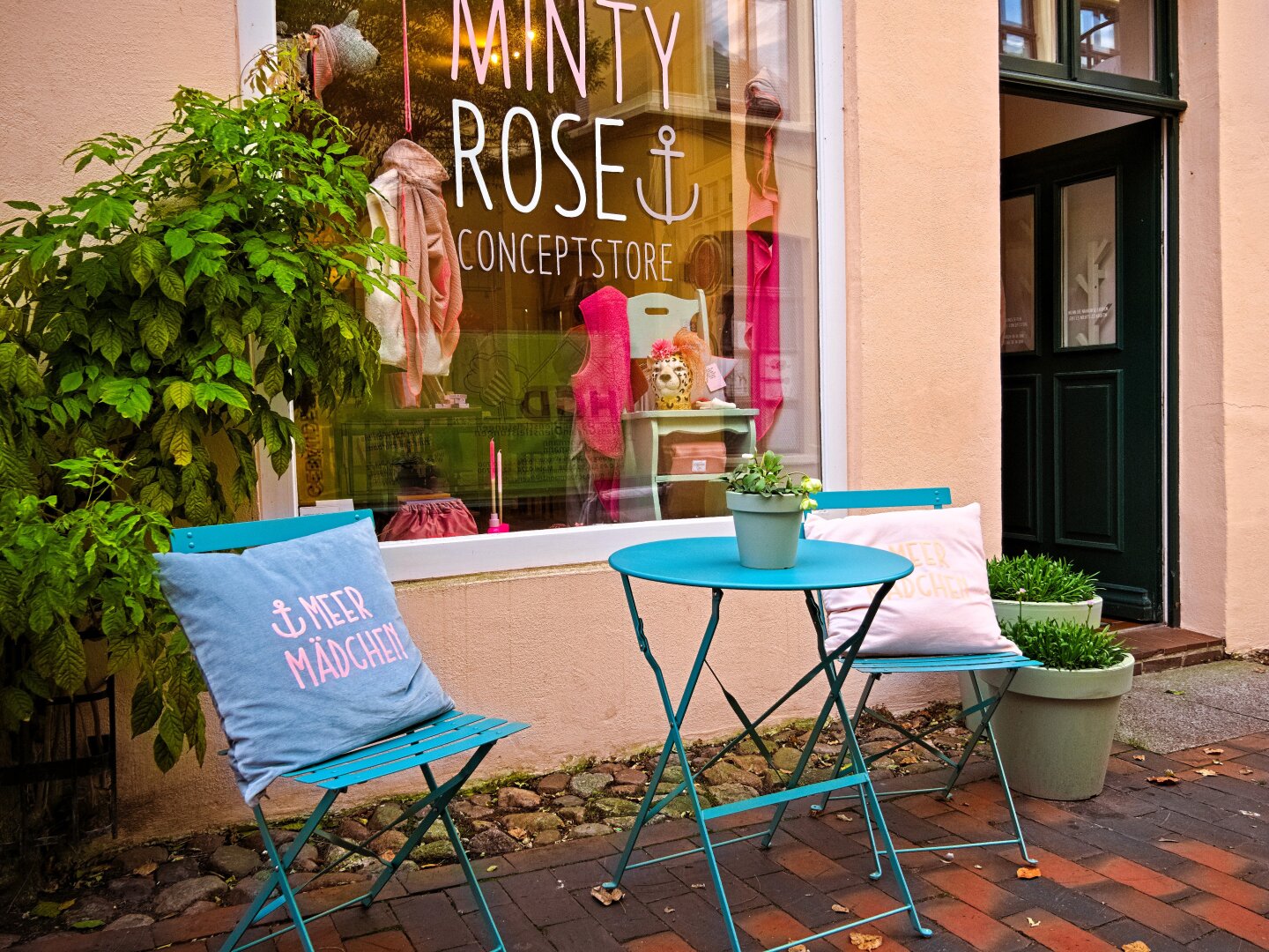 In front of a shop called ‘Minty Rose’ are two folding chairs and a table. They are painted in a pretty shade of blue. On each chair is a cushion with the words ‘Meer Mädchen' (Sea girl) written on it. The shop window is decorated with clothing and accessories, all in shades of pink and brown. This matches the front of the building, which is light in colour with a pinkish-brown tinge. Next to the shop window is an open green door. There are also plants in front of the window. I think it looks inviting.